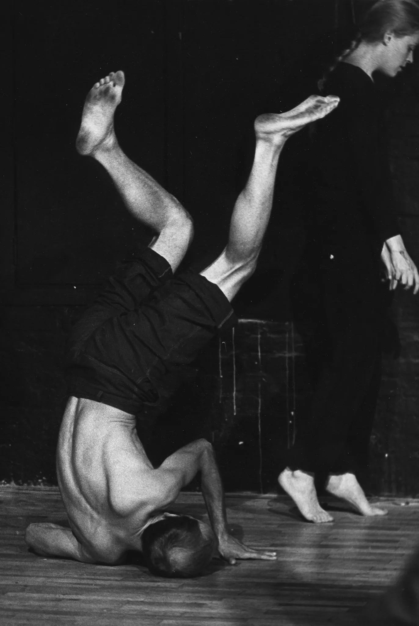 Aktor stoi na przekrzywionej głowie podpierając się rękami. Jego ciało jest wygięte. Ustawiony jest plecami w stronę widza. Po prawej stronie aktorka stoi na palcach. Jest ubrana na czarno i jest boso. Jej włosy są splecione w warkocz.