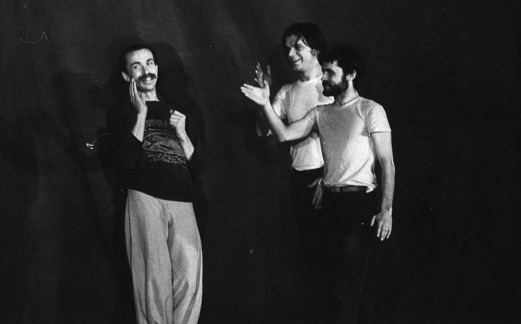 Trzech aktorów. Aktor po lewej stronie patrzy w przestrzeń i uśmiecha się. Ubrany jest w jasne spodnie i czarną bluzę z nadrukowanym wzorem. Po prawej stronie dwóch aktorów sięga w jego stronę ręką. Również się uśmiechają. Ubrani są w czarne spodnie i białe podkoszulki.