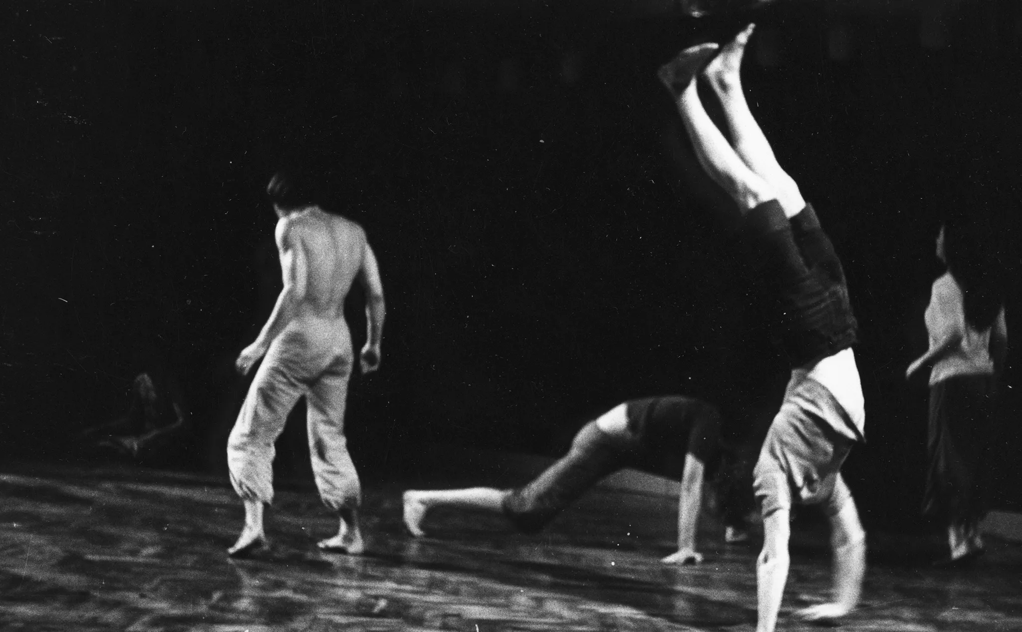 Po lewej stronie, zwrócony tyłem do widza stoi aktor. Ubrany jest w jasne spodnie, ma nagi, muskularny tors. Jest boso. W centrum dwoje aktorów wykonuje akrobatyczne pozy. Jeden stoi na rękach, drugi wygląda, jakby szykował się do robienia pompek. Po prawej stronie na palach stoi aktorka.