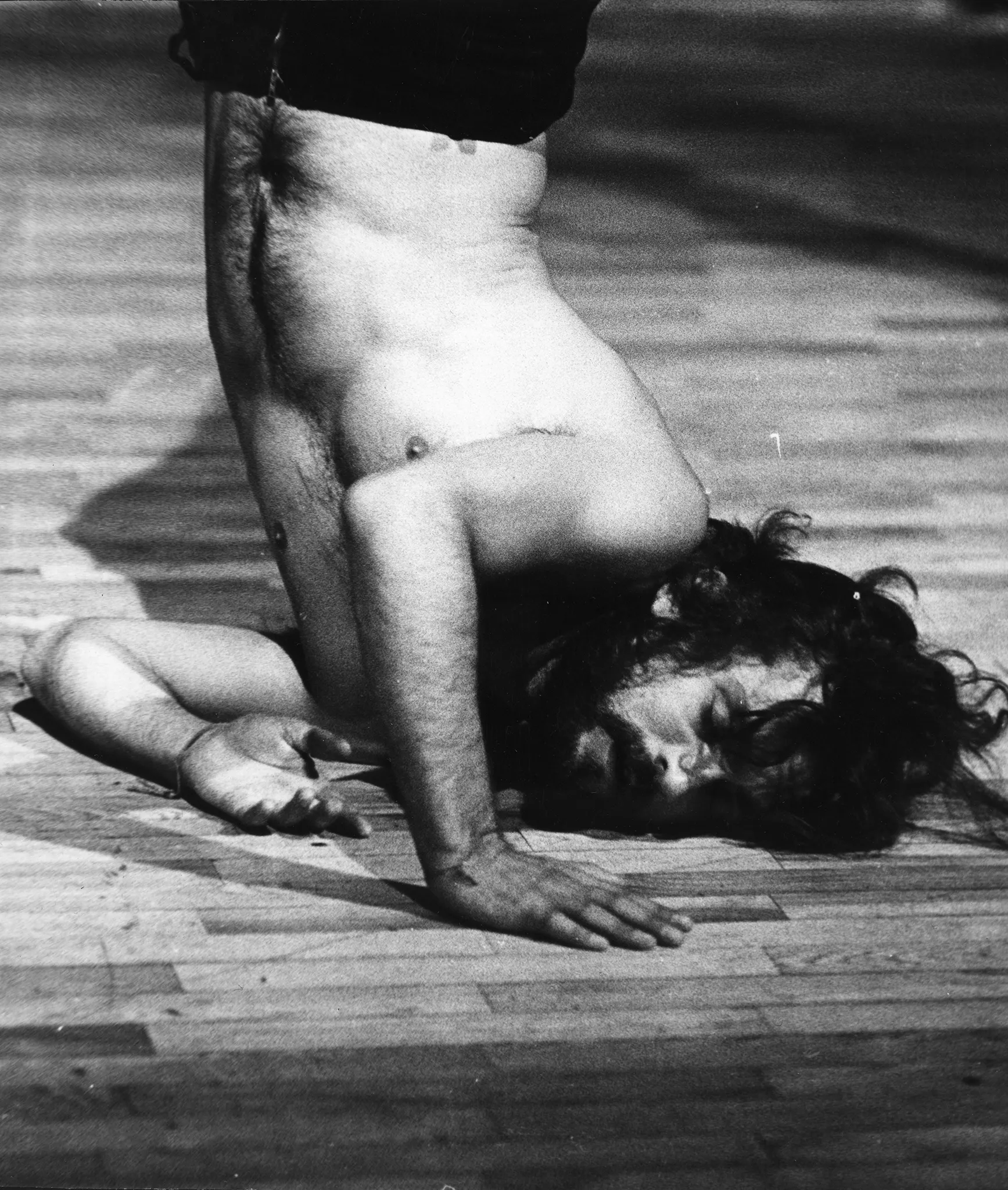 Aktor stoi na przekrywionej głowie podpierając się rękami. Ustawiony jest twarzą w stronę widza. Ma długie włosy i gęstą brodę. Jego tors jest nagi. Aktor widoczny jest tylko do pasa.