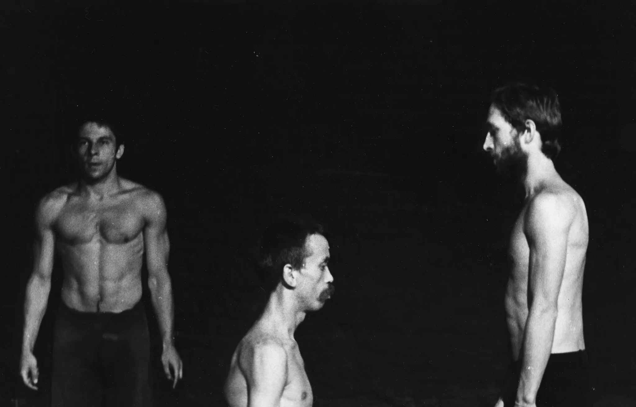 Po lewej stronie, zwrócony twarzą do widza, stoi aktor widoczny od pasa w górę. Ma ciemne spodnie i nagi, muskarlny tors. Po prawej stronie, zwróceni profilem do widza, stoją dwaj aktorzy. Są zwróceni do siebie twarzami. Aktor po lewej stronie wygląda, jakby klęczał przed aktorem po prawej. Obaj mają nagie torsy.