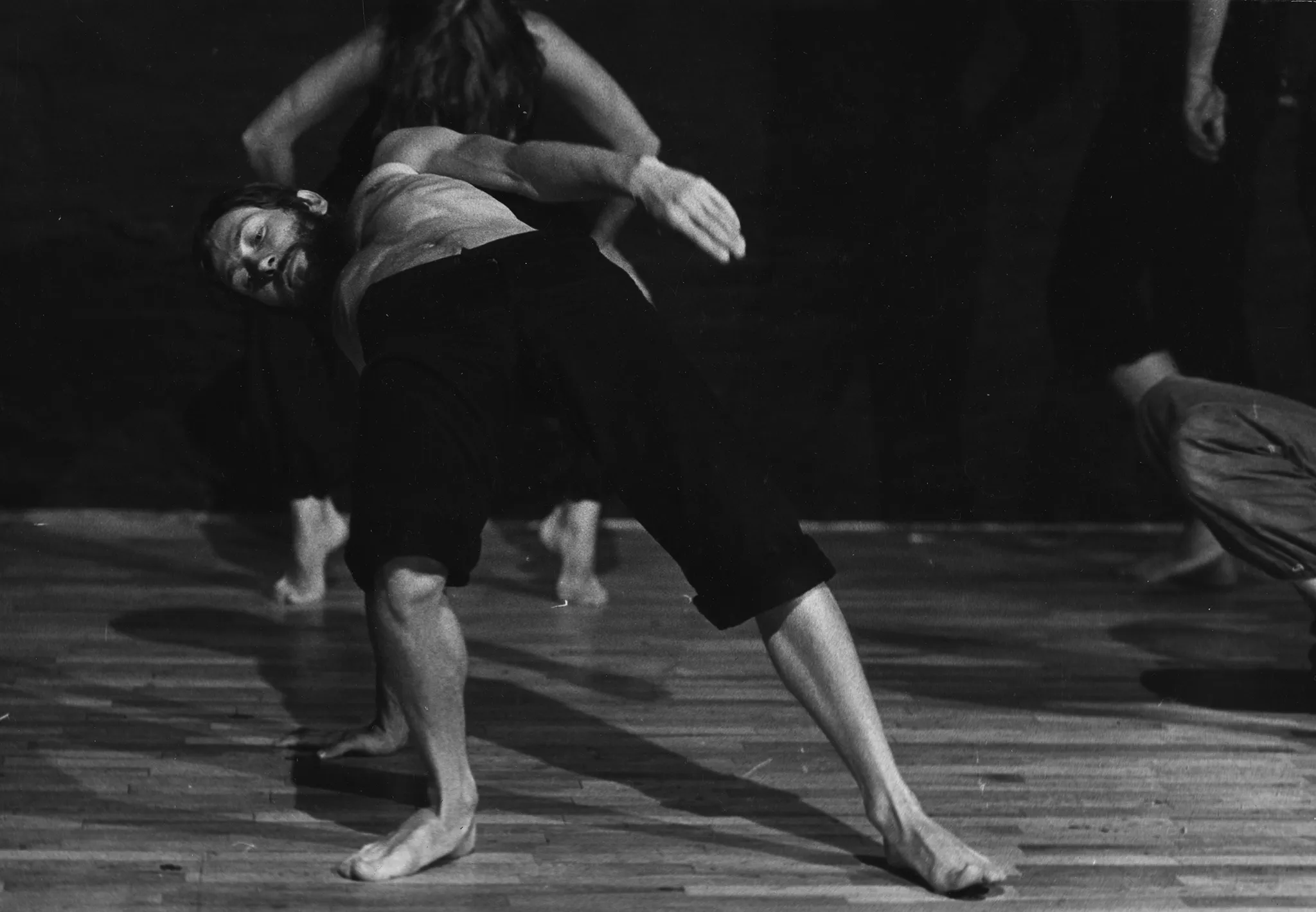 Aktor, ustawiony brzuchem do góry, podpiera się jedną ręką o podłogę. Ubrany jest w czarne spodenki, ma nagi tors. Jest boso. Patrzy w podłogę. Za jego plecami słabo widoczna aktorka stoi na palcach i zwisa głową w dół.