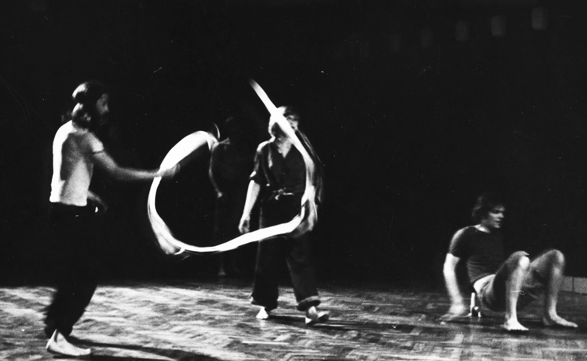 Po lewej stronie stoi aktor ubrany w ciemne spodnie i jasny podkoszulek. Jest boso. Macha wstążką gimnastyczną. Obok niego stoi przysłonięta wstążką aktorka. Ubrana jest na czarno, jest boso. Po prawej stronie, zwrócony brzuchem do góry, podpiera się na rękach aktor. Ubrany jest w czarny podkoszulek. Jest boso.