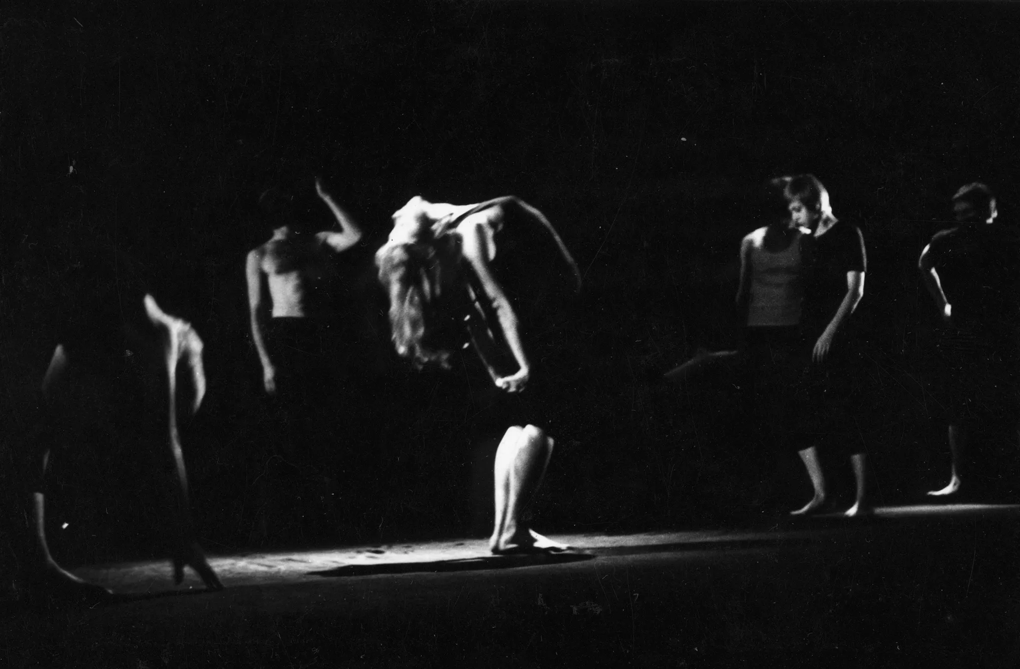 Grupa aktorów i aktorek wykonuje oddzielne działania. W centrum stoi aktorka wyginająca ciało do tyłu. Jest ubrana na czarno. Jest boso. Jej długie włosy zwisają do tyłu.