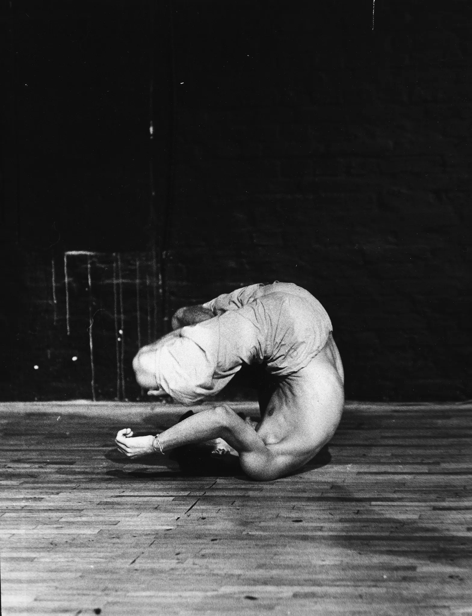 Aktor ubrany w jasne spodenki. Jest boso i ma nagi tors. Wygląda, jakby miał się przeturlać na podłodze.