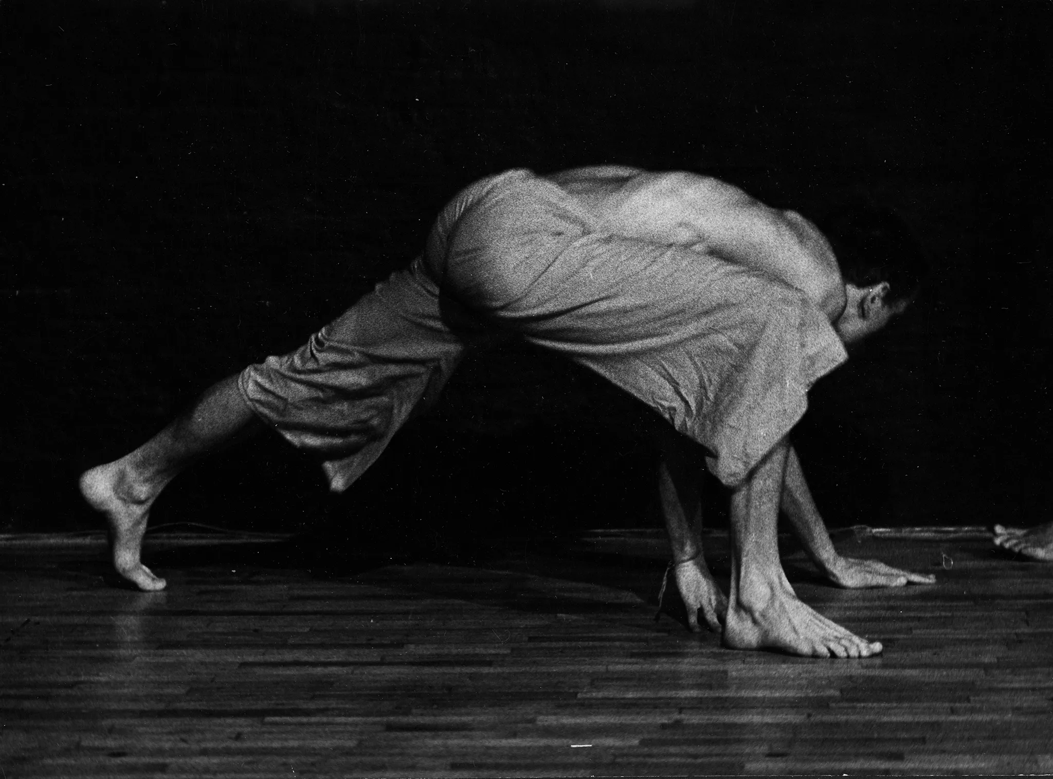 Aktor ubrany w jasne spodenki. Jest boso i ma nagi tors. Wygląda, jakby miał się przeturlać na podłodze.