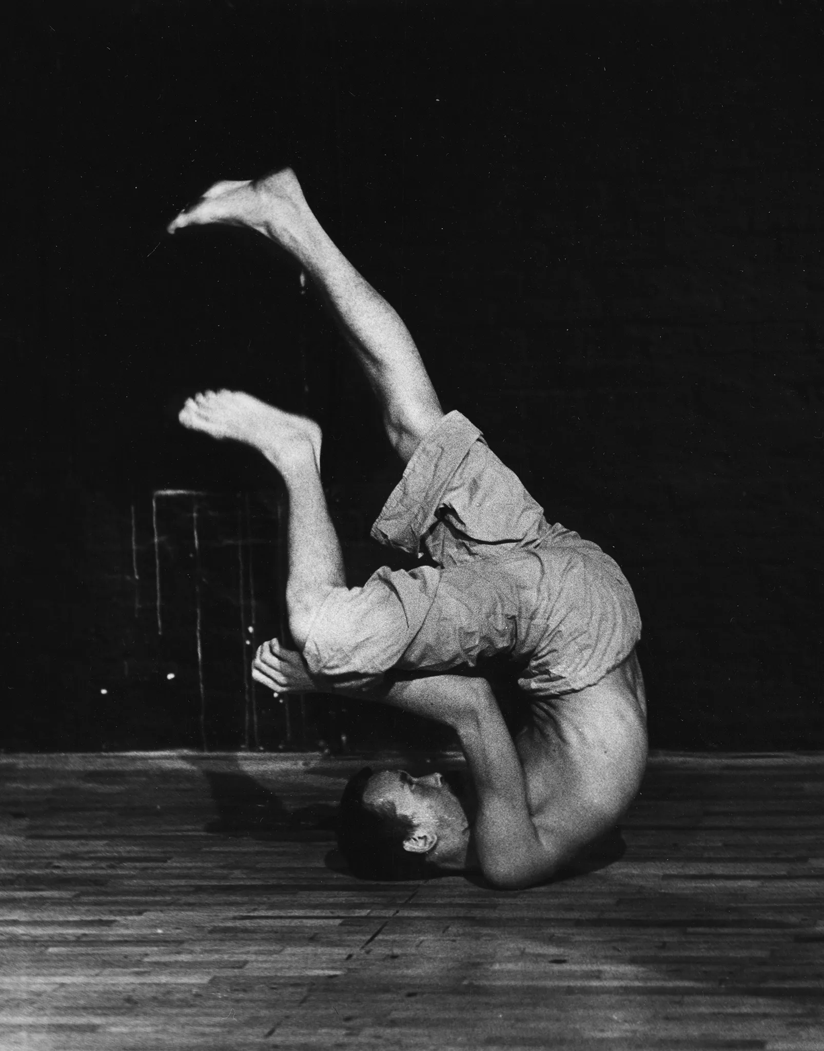 Aktor ubrany w jasne spodenki. Jest boso i ma nagi tors. Wygląda, jakby miał się przeturlać na podłodze.