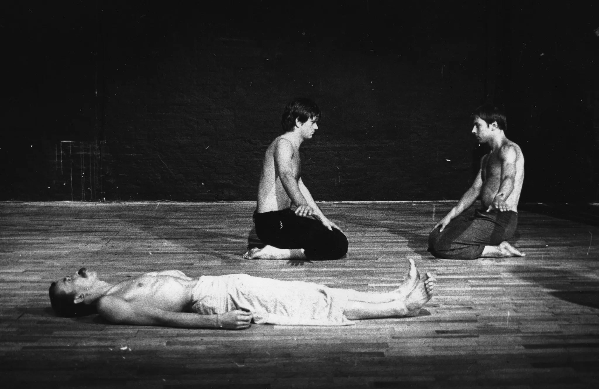 Na plecach leży aktor. Ubrany jest w jasne spodenki, ma nagi tors. Patrzy w sufit. Za nim klęczy dwóch aktorów. Ubrani są w ciemne spodenki, mają nagie torsy. Klęczą naprzeciwko siebie i patrzą na siebie nawzajem. Obaj mają jedną rękę wyciągniętą do boku.