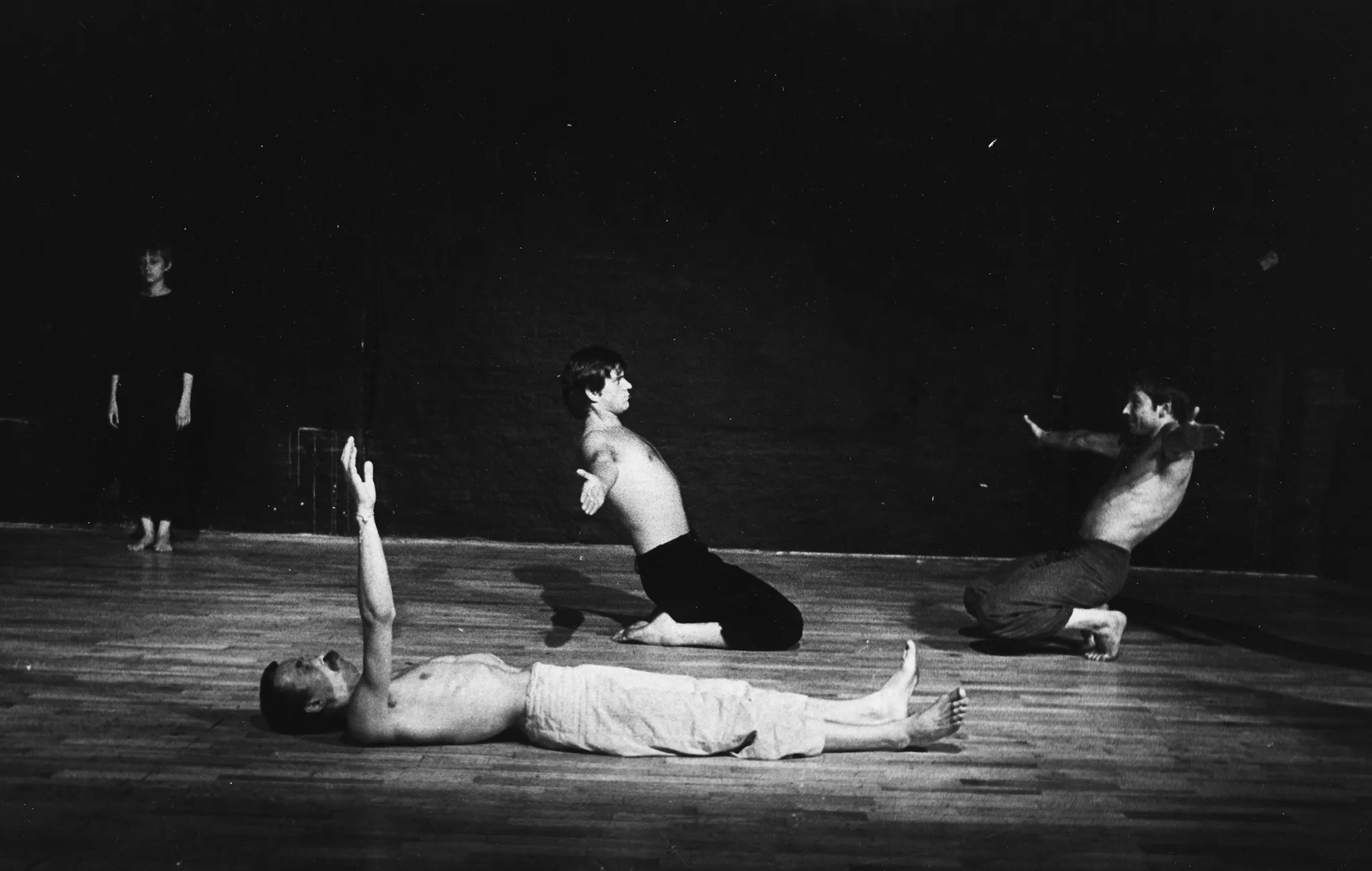 Po lewej stronie stoi zwrócona w stronę widza aktorka. Ubrana jest w czarne spodenki i czarną podkoszulkę. Jest boso. W centrum, na pierwszym planie na plecach leży aktor. Ubrany jest w jasne spodenki, ma nagi tors. Patrzy w sufit i wyciągata przed siebie jedną rękę. Za nim klęczy dwóch aktorów. Ubrani są w ciemne spodenki, mają nagie torsy. Klęczą naprzeciwko siebie i patrzą na siebie nawzajem. Mają ręce wyciągnięte na boki.