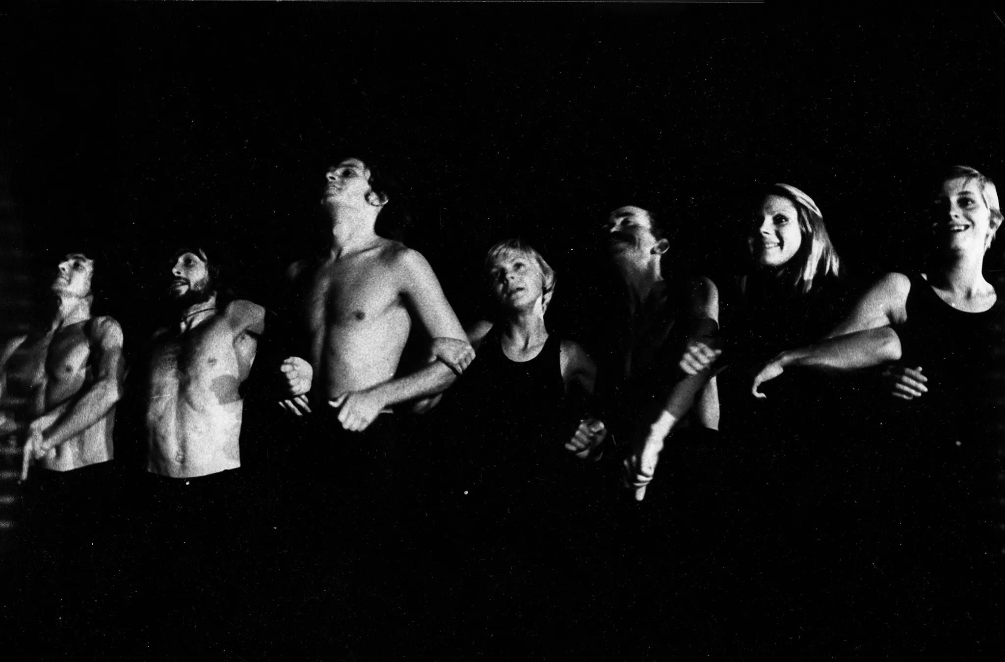 Grupa aktorów i aktorek stojących w szeregu. Aktorzy mają nagie torsy, aktorki ubrane są w czarne koszulki na ramiączka. Patrzą przed siebie, chwytają się pod rękę, uśmiechają się. Aktorzy napinają mięśnie.