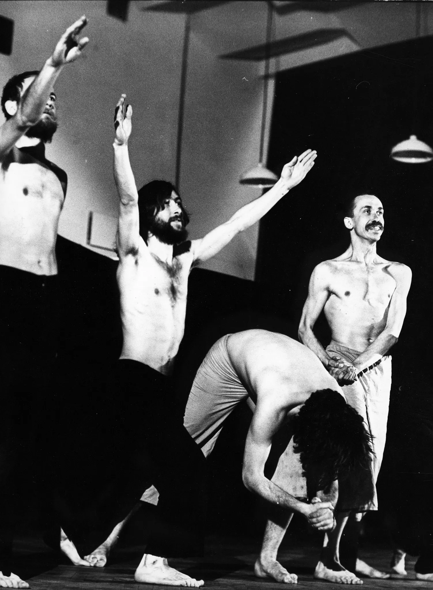 Czterech aktorów stoi w szeregu. Dwóch aktorów po lewej stronie ubranych jest w czarne spodenki. Mają uniesione ręce. Obok nich, w środku, stoi aktor zwisający głową ku podłodze. Po prawej stronie stoi aktor ubrany w jasne spodenki. Napina mięśnie i uśmiecha się. Wszyscy mają nagie torsy i są boso. Z sufitu zwisają lampy z szerokimi kloszami.