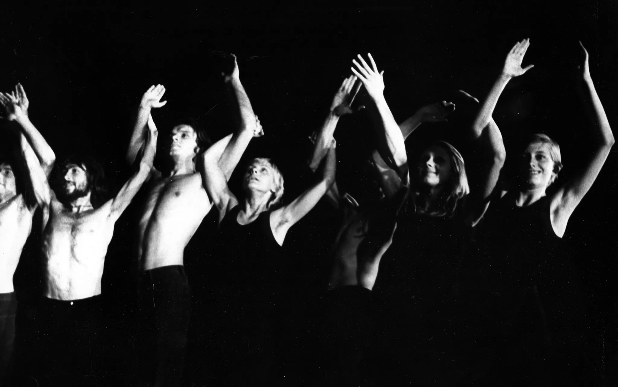 Grupa aktorów i aktorek stojących w szeregu. Aktorzy mają nagie torsy, aktorki ubrane są w czarne koszulki na ramiączka. Patrzą przed siebie, uśmiechają się. Mają ręce uniesiono wysoko w górę.