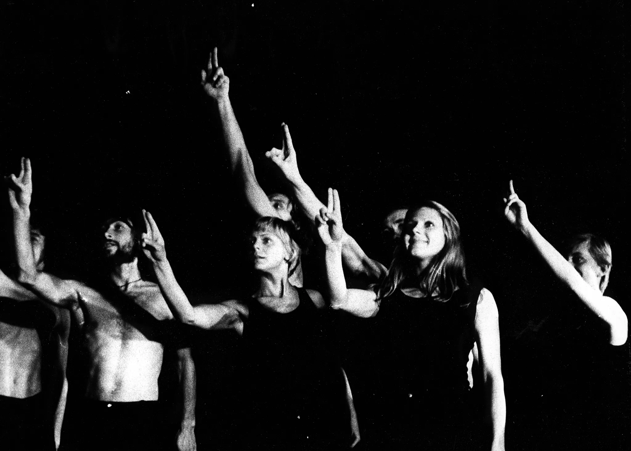 Grupa aktorów i aktorek stojących w szeregu. Aktorzy mają nagie torsy, aktorki ubrane są w czarne koszulki na ramiączka. Patrzą przed siebie. Wszyscy mają uniesioną rękę i wyprostowanymi dwoma palcami, jakby zgłaszali się do odpowiedzi.