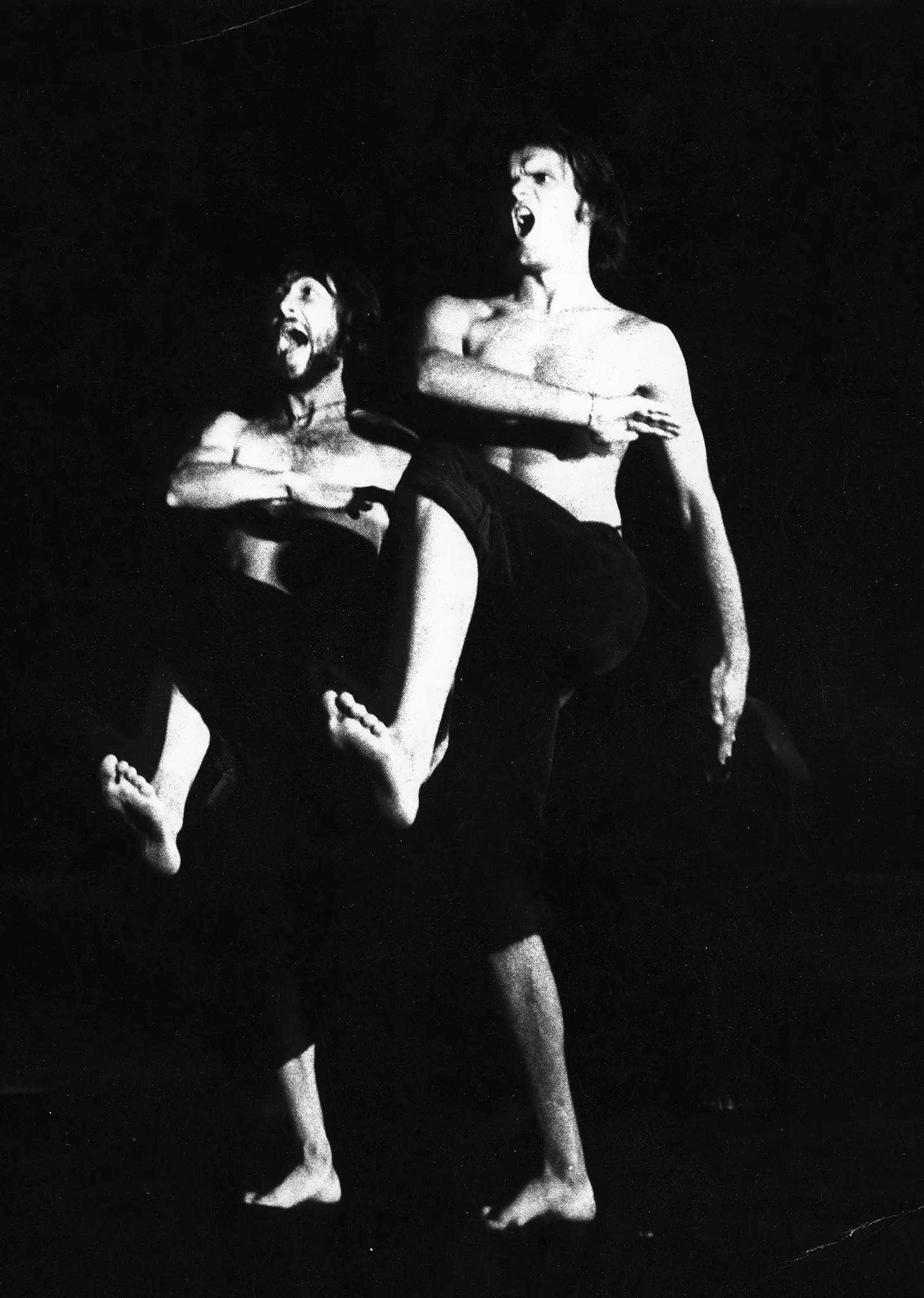 Dwóch aktorów w szeregu maszeruje energicznie przed siebie. Wyglądają, jakby krzyczeli. Ubrani są w czarne spodenki, mają nagie torsy. Są boso.