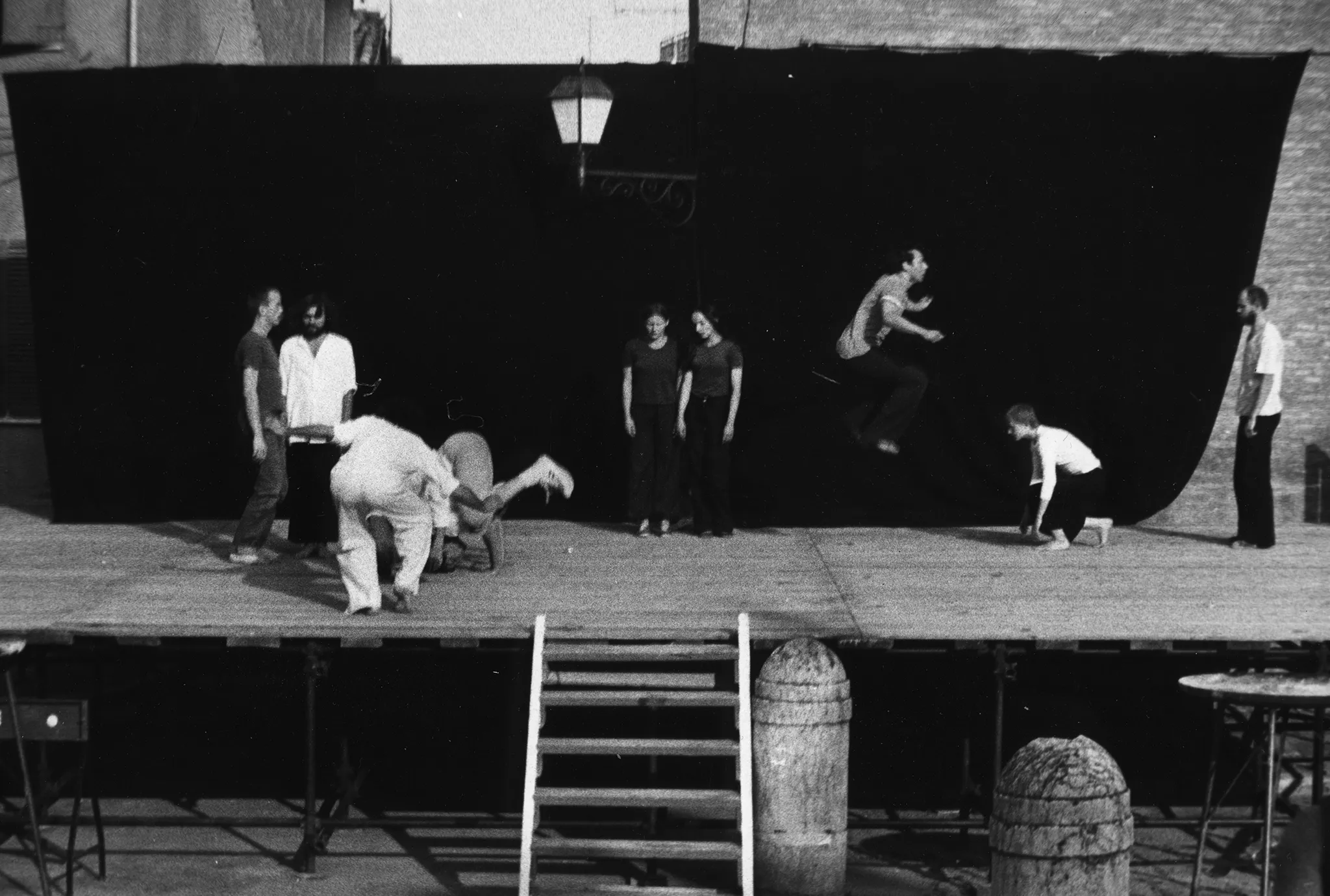 Scena plenerowa. Na scenę prowadzą drewniane schody. Z tyłu sceny ściana z czarnej płachty. Pośrodku, zza płachty wystaje latarnia. Na scenie stoją aktorzy i aktorki w różnych działaniach. Turlają się, skaczą i stoją w miejscu. Wyglądają, jakby mieli na sobie prywatne ubrania. Wspólnie tworzą choreograficzną kompozycją.