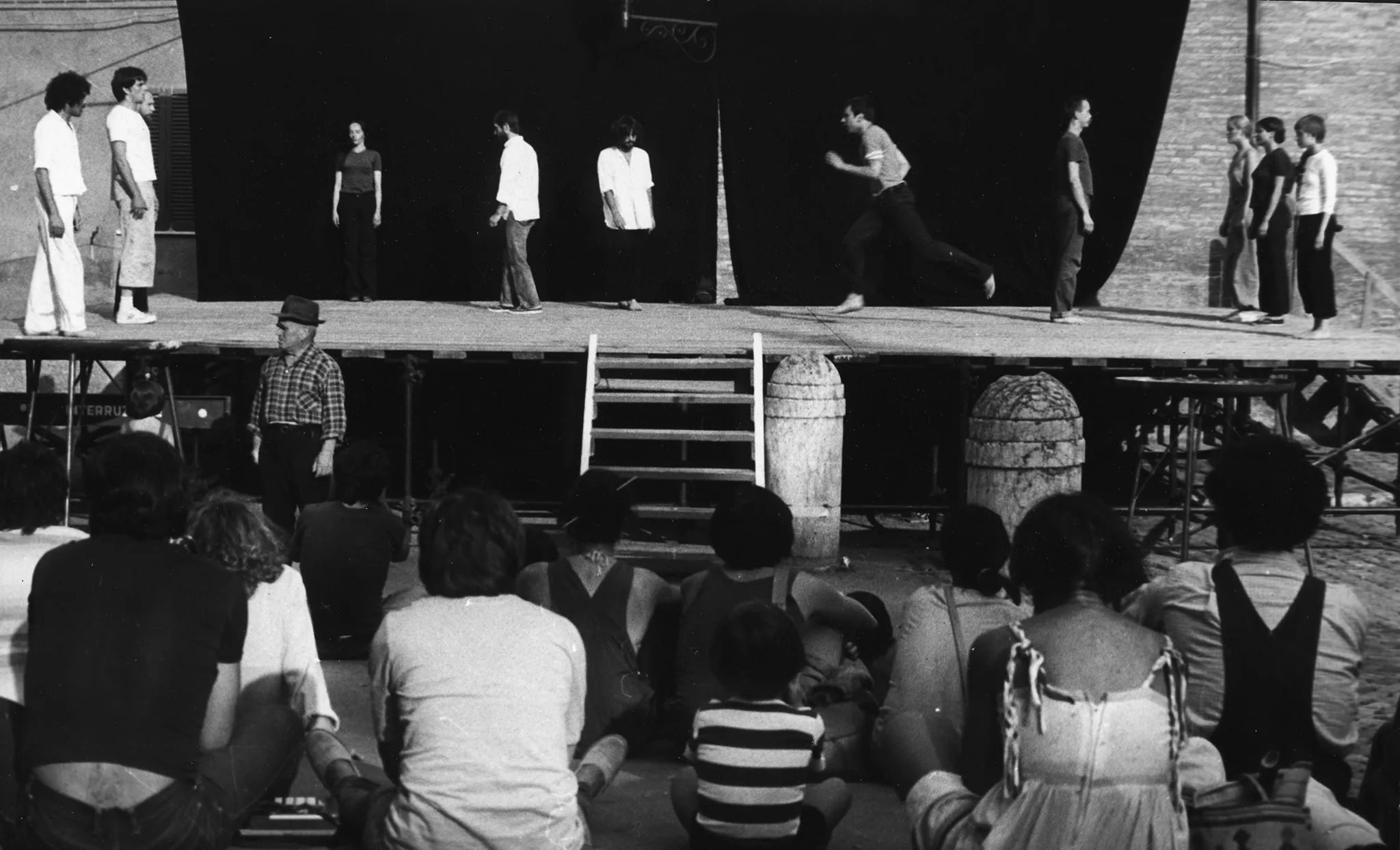 Scena plenerowa. Na pierwszym planie odwróceni plecami widzowie patrzą na scenę. Na scenę prowadzą drewniane schody. Z tyłu sceny ściana z czarnej płachty. Na scenie stoją aktorzy i aktorki w różnych działaniach. Wyglądają, jakby mieli na sobie prywatne ubrania. Wspólnie tworzą choreograficzną kompozycję. Aktorzy i aktorki po bokach sceny są statyczni. Aktorzy w centrum ruszają się dynamicznie. Na dalszym planie, za sceną widać budynki.