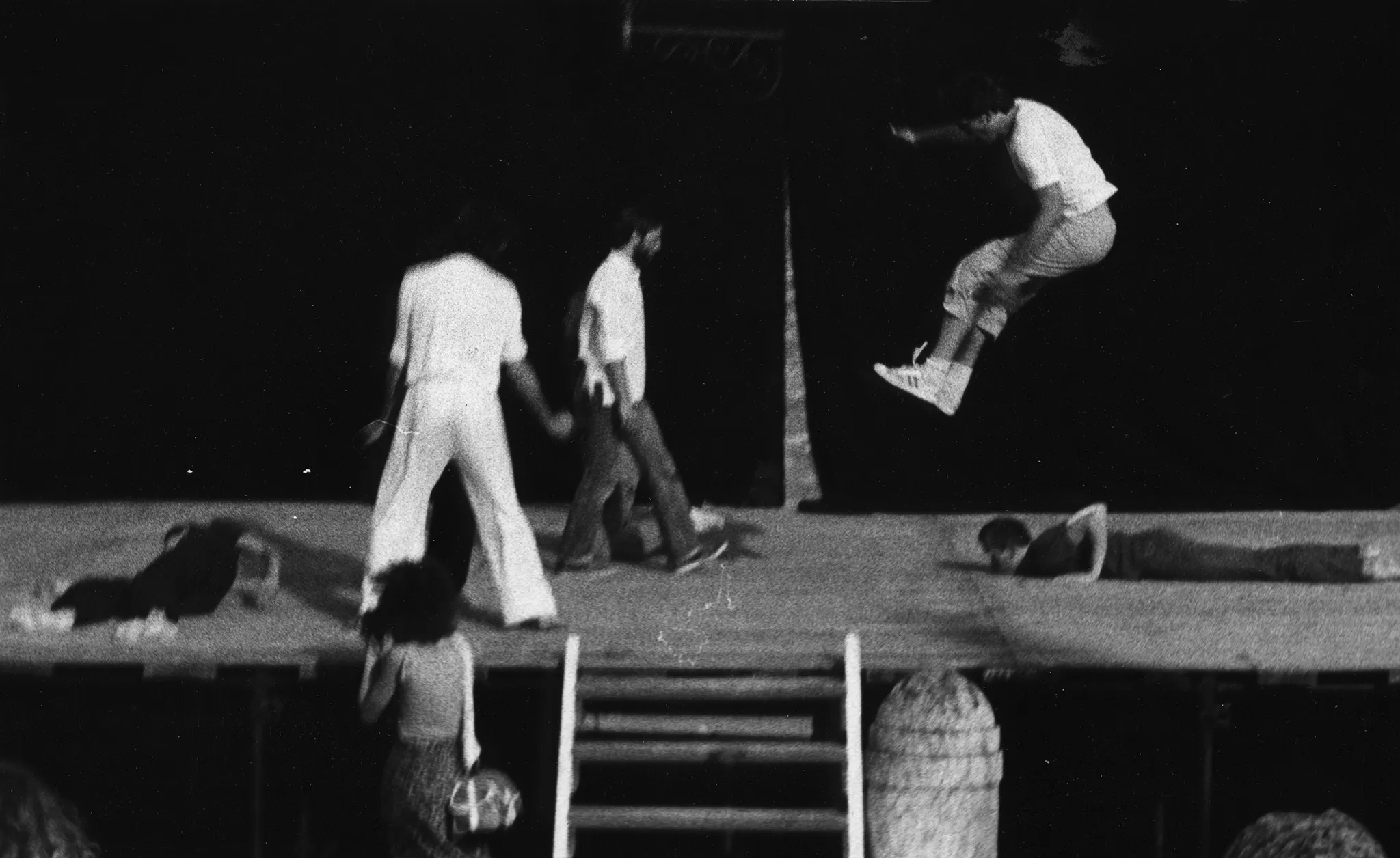 Scena plenerowa. Na scenę prowadzą drewniane schody. Z tyłu sceny ściana z czarnej płachty. Pod sceną stoi widzka. Na scenie, po lewej stronie na plecach leży aktorka ubrana na czarno. Obok niej stoi zwrócony plecami do widza aktor ubrany na biało. Obok niego przechodzi zwrócony profilem do widza aktor ubrany w jeansy i białą koszulę. Z prawej strony sceny bardzo wysoko skacze aktor. Ubrany jest w jasne buty sportowe, jasne spodnie i jasną podkoszulkę. Za nim na brzuchu leży aktor ubrany na czarno.