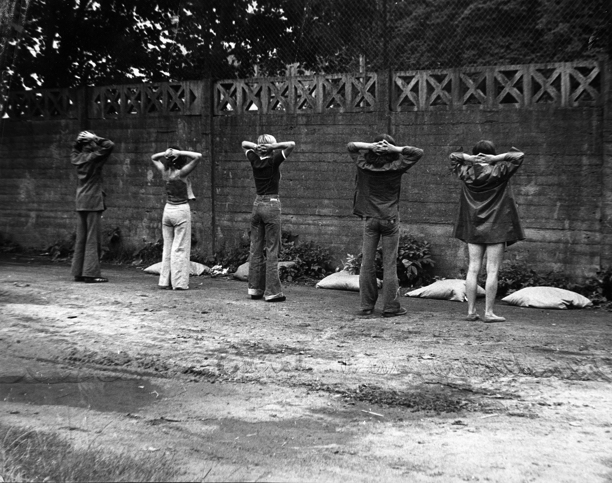 Piaszczysta droga z kałużami. Pod betonowym płotem stoją odwróceni plecami do widza aktorzy. Mają ręce założone z tyłu głowy. Między płotem a aktorami leżą białe worki.