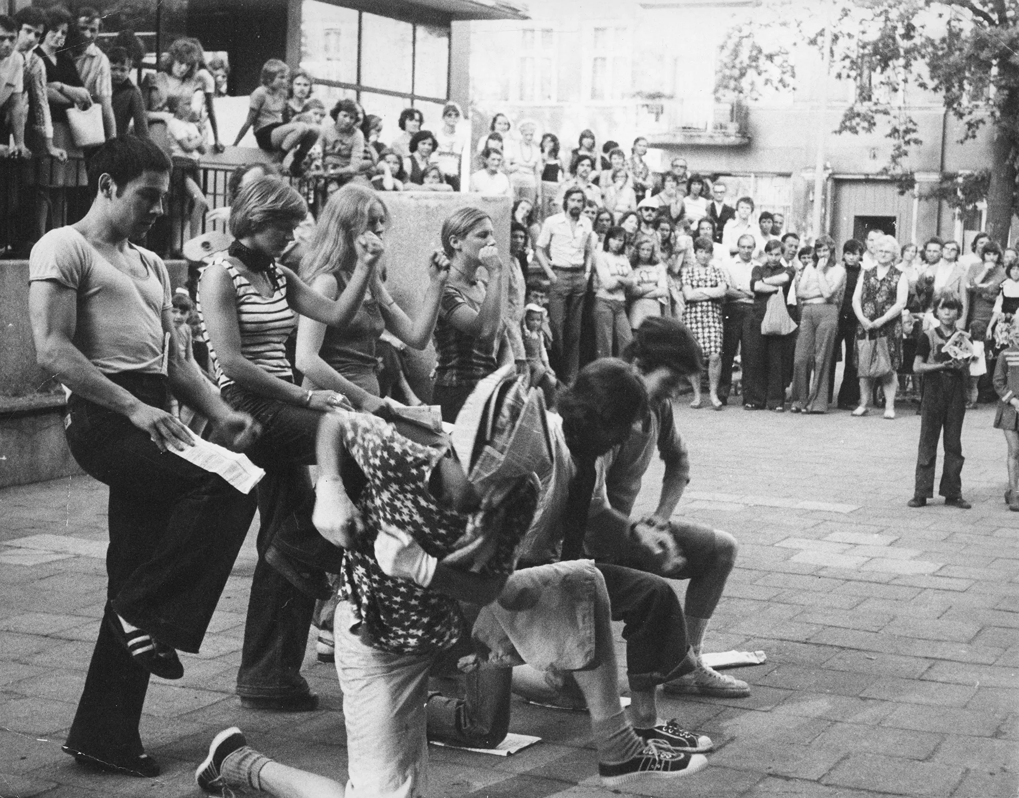 Przestrzeń miejska biędzy budynkami. Aktorzy i aktorki w dwóch szeregach. Aktorzy w pierwszym szeregu klęczą na jednej nodze. Aktor i aktorki w drugim szeregu stoją z jedną nogą uniesioną. Ich ubrania wyglądają na prywatne. Są ubrani w trampki, jeansy i koszulki. Działaniu aktorów przygląda się duża grupa otaczających ich widzów.