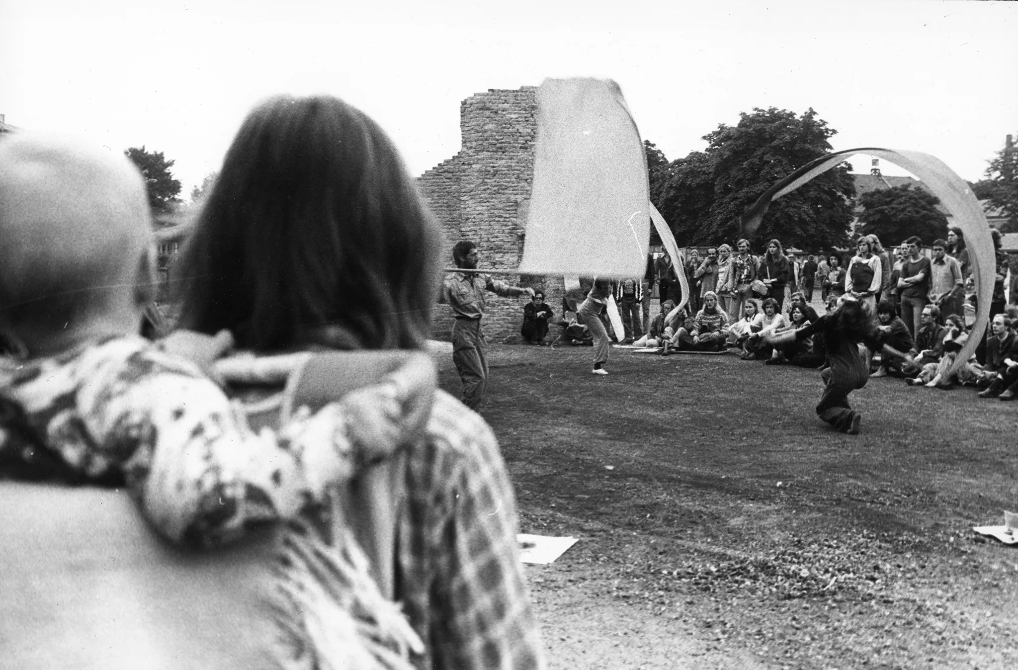 Plener, ruiny jakiegoś budynku. Troje aktorów energicznie macha białymi flagami i wstęgami. W tle siedzą i stoją widzowie, którzy oglądają aktorów. Na pierwszym planie widać plecy widzki trzymającej niemowlę na plecach.