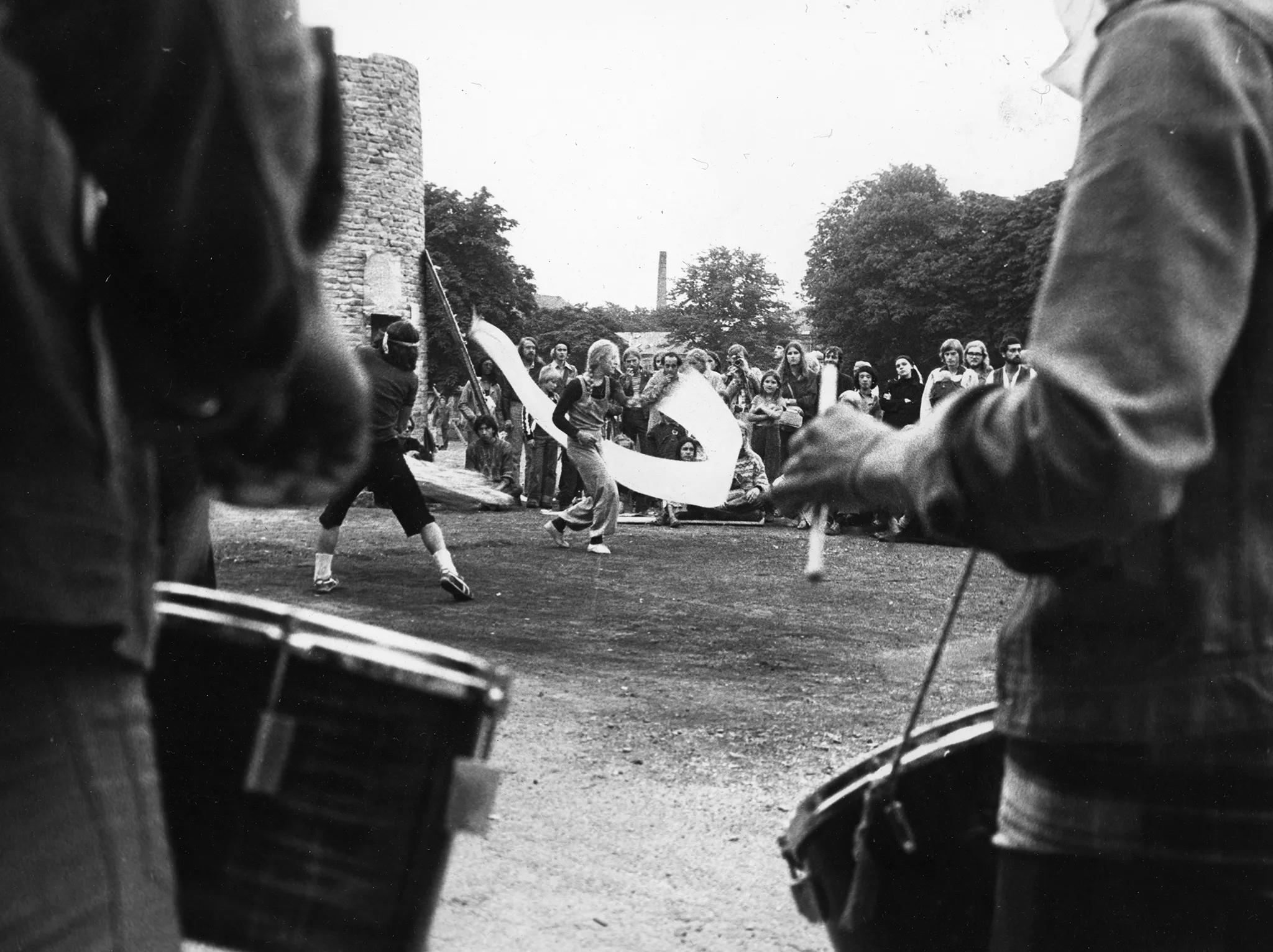 Plener, ruiny jakiegoś budynku. Grupa aktorów i aktorek macha białymi wstęgami. W tle siedzą i stoją widzowie, którzy oglądają aktorów. Na pierwszym planie obróceni plecami do widza dwaj mężczyźni grający na bębnach.