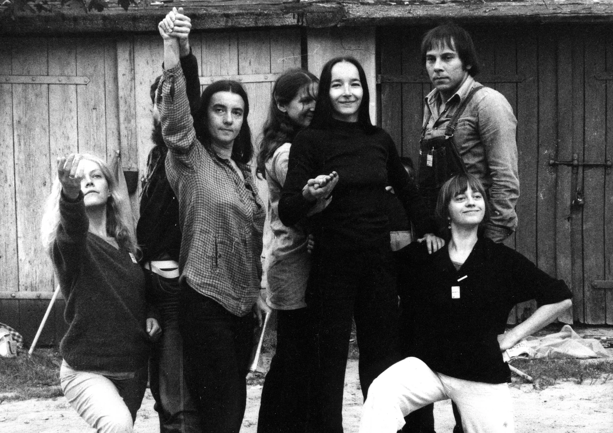 Plener. Grupa aktorek i jeden aktor pozuje do zdjęcia. Dwie aktorki po bokach klęczą, są uśmiechnięte. Cztery aktorki w środku trzymają się za ręce, są uśmiechnięte. Aktor stoi nad klęczącą aktorką po prawej stronie. Lekko się uśmiecha. W tle widać budynek z kilkoma drewnianymi drzwiami.