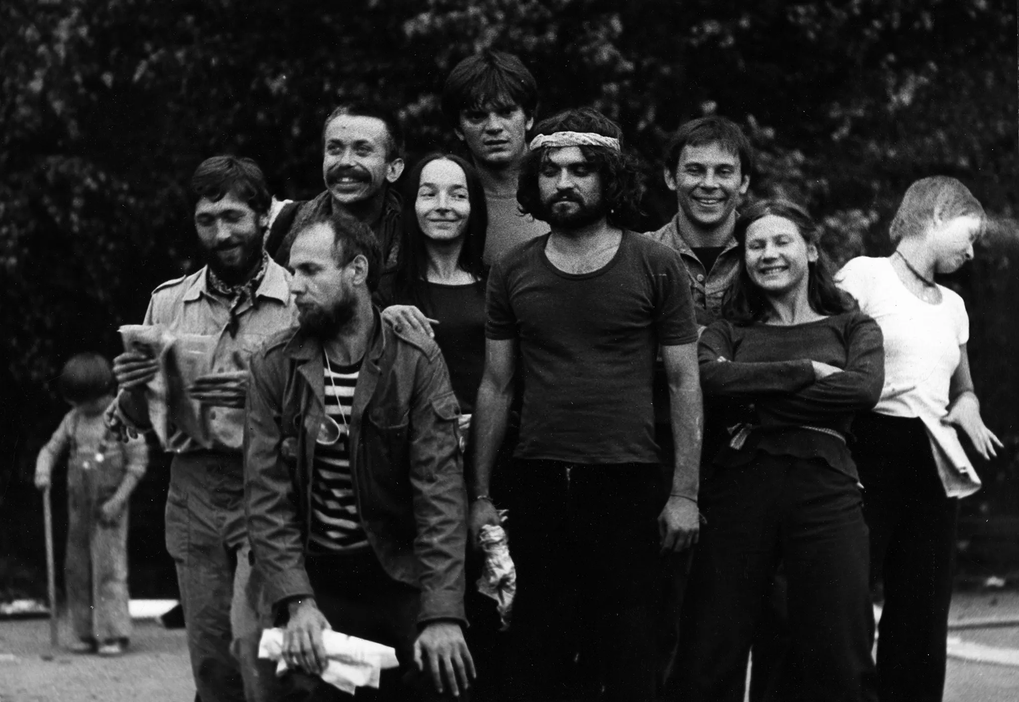 Plener. Grupa aktorów i aktorek pozuje do zdjęcia. Stoją bardzo blisko siebie, uśmiechają się. Wszyscy mają ciemne ubrania poza wyróżniającą się aktorką z prawej strony. Aktorka ubrana jest w białą podkoszulkę i patrzy w bok. Na dalszym planie po lewej stronie stoi dziecko bawiące się kijem. W tle widać drzewa.