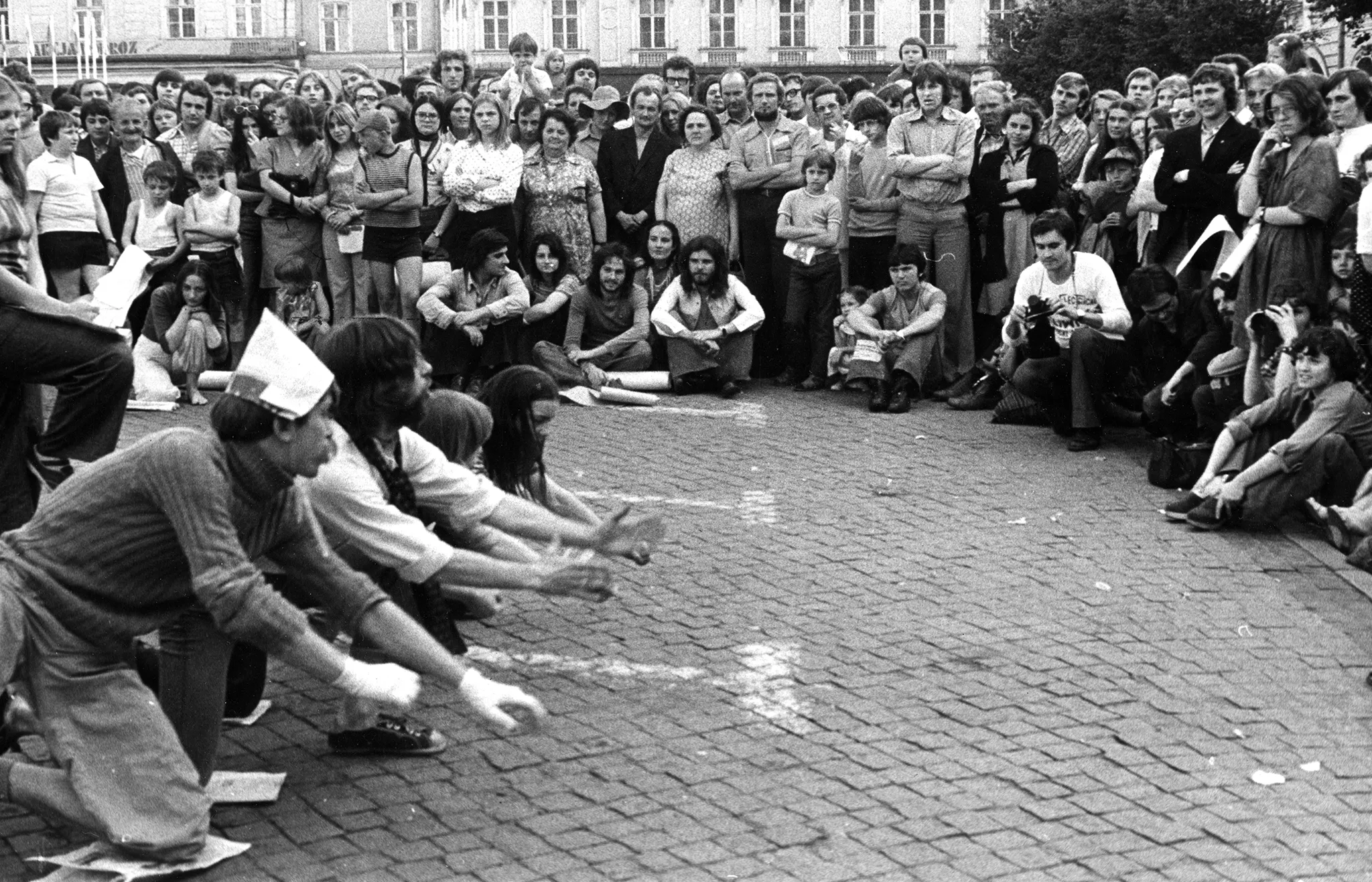 Grupa aktorów i aktorek na brukowanej ulicy. Każdy z nich klęczy na jednej nodze. Pod kolanami mają gazety. Wyciągają ręce w stronę dużej grupy widzów stojących dookoła. W tle widać budynki mieszkalne.