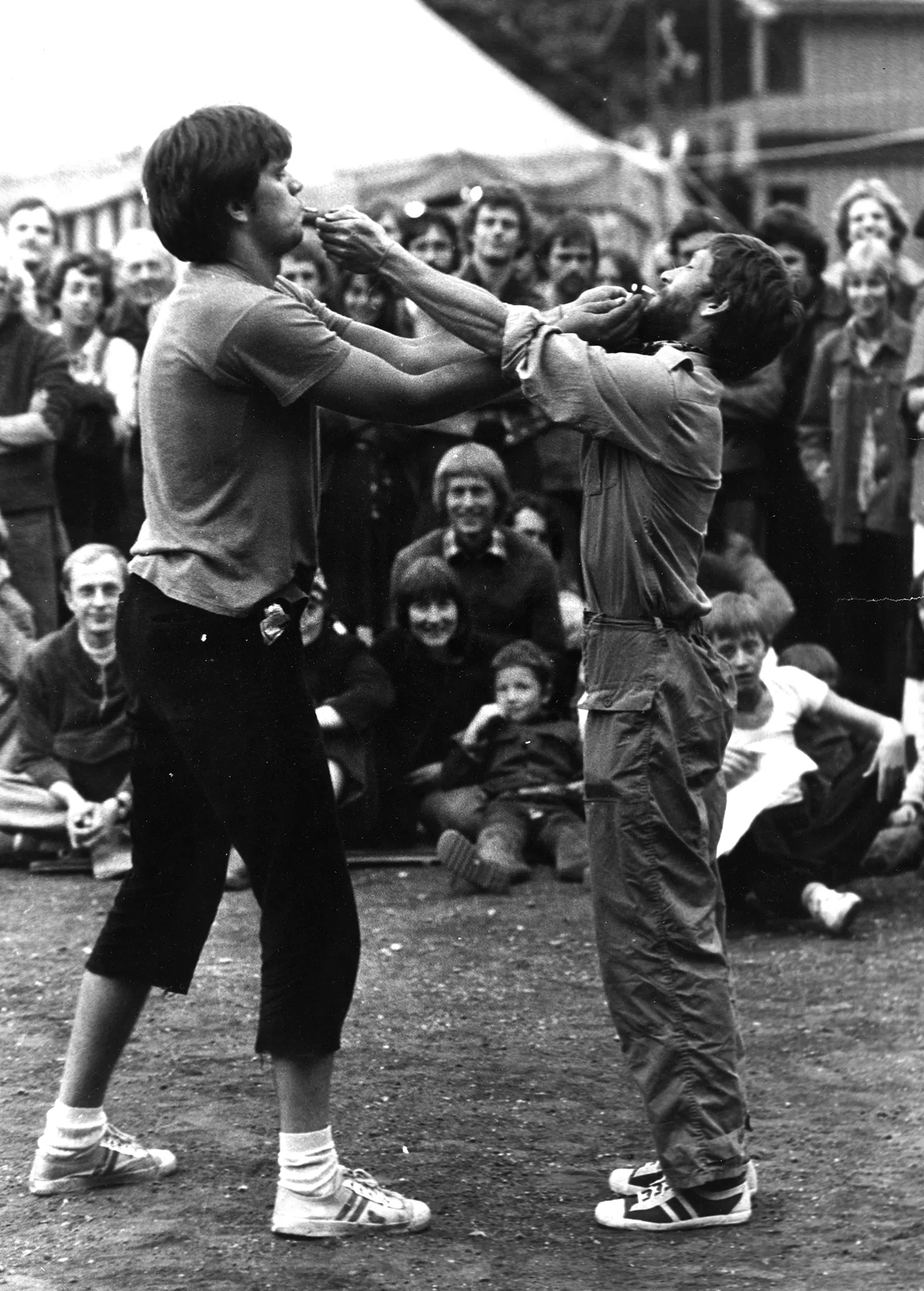 Dwóch aktorów stoi naprzeciwko siebie. Są ubrani w trampki, spodnie i podkoszulki. Podpalają sobie wzajemnie papierosy, które trzymają w ustach. Na drugim planie widać grupę widzów oglądających aktorów.