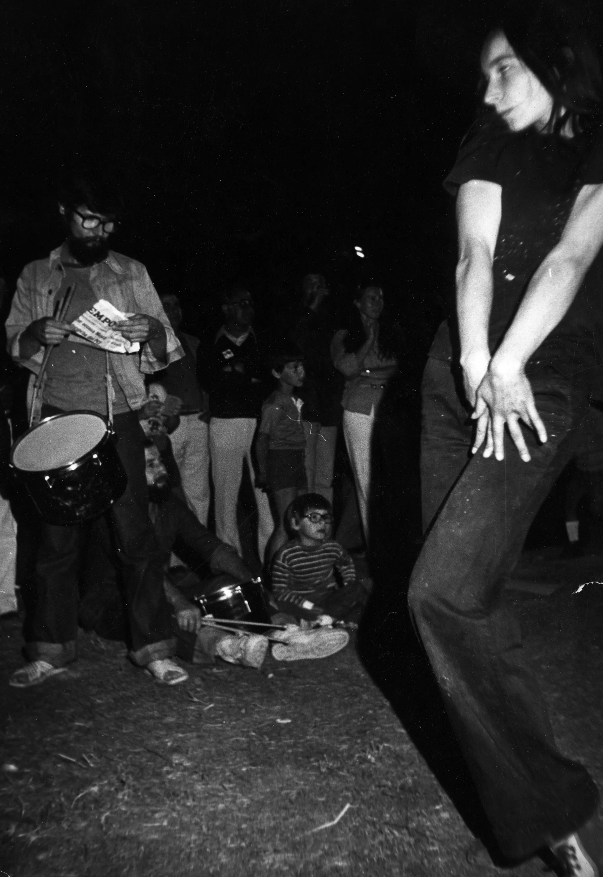 Na pierwszym planie stoi zwrócona twarzą do widza aktorka ubrana na czarno. AKtorka lekko ugina nogę. Na ugiętej nodze trzyma obie dłonie. Na drugim planie stoi aktor z bębnem zawieszonym na szyi. W tle stoi grupa widzów.