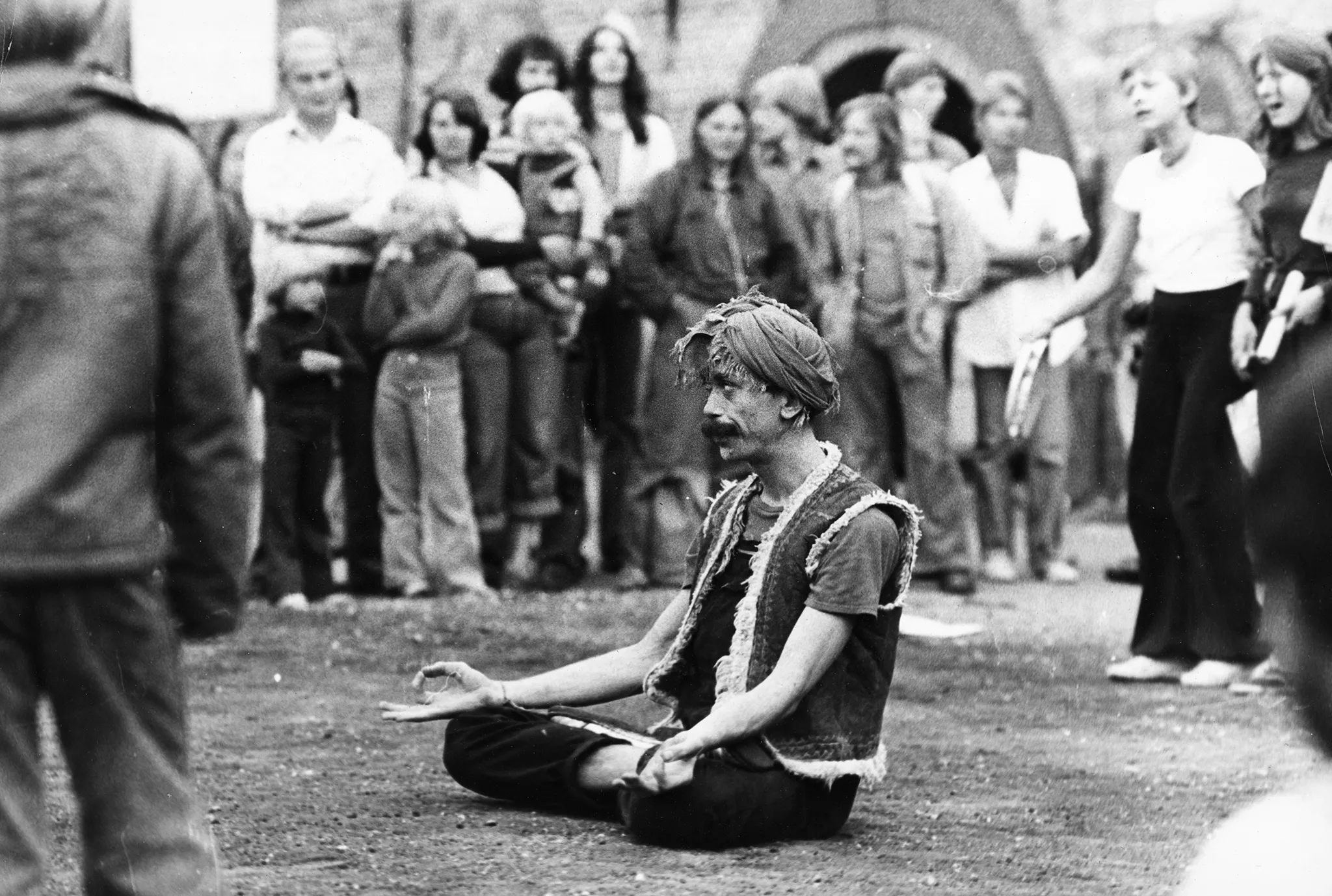 W centrum siedzi aktor w siadzie skrzyżnym. Wygląda, jakby medytował. Trzyma zgięte ręce po bokach. Na głowie ma zawiązaną chustę. Dookoła niego stoi grupa widzów.