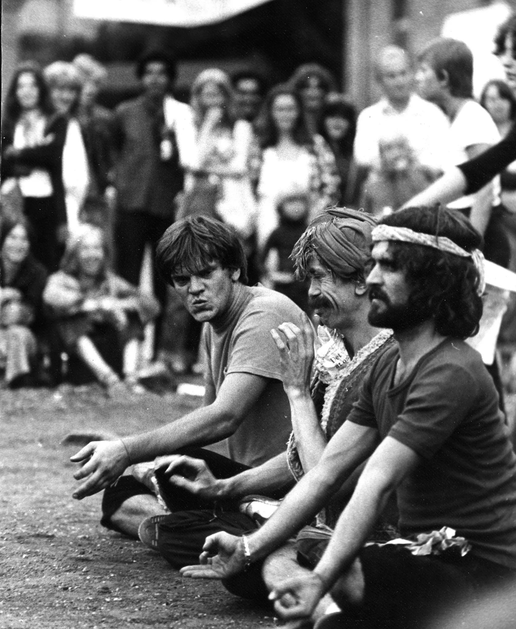 Trzech aktroów siedzi w szeregu w siadzie skrzyżnym. Mają zabawny wyraz twarzy. Na dalszym planie siedzą i stoją widzowie. Śmieją się.