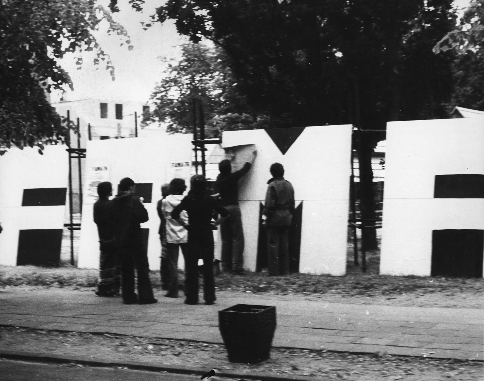 Przestrzeń miejska. Na chodniku stoi odwrócona plecami do widza grupa aktorów. Ustawiają na trawniku wielki biały napis "FAMA". W tle widać drzewa i budynki.