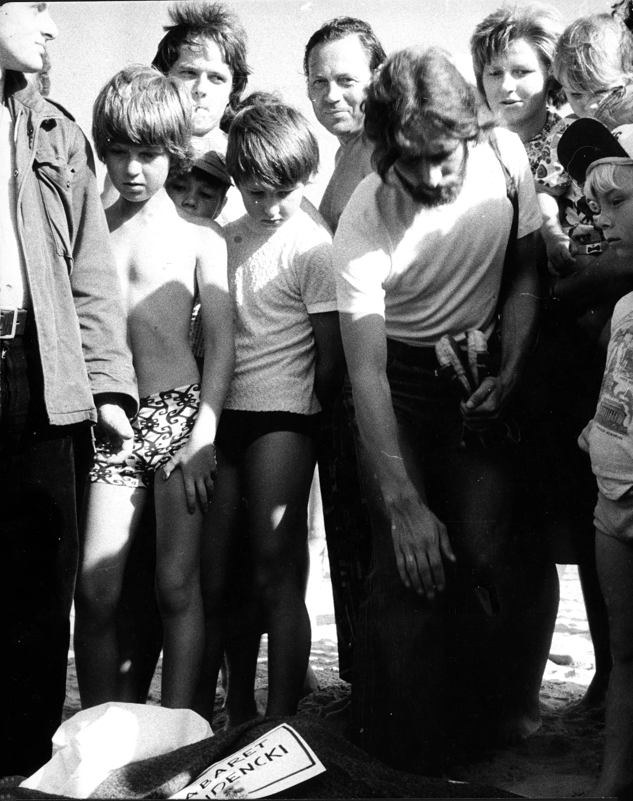 Grupa osób, dzieci i dorosłych, stoi na plaży. Dzieci ubrane są w majtki i podkoszulki, dorośli mają jeansy i podkoszulki. Niektórzy patrzą w stronę widza i uśmiechają się. Na ziemi leży tabliczka przypominająca nekrolog.