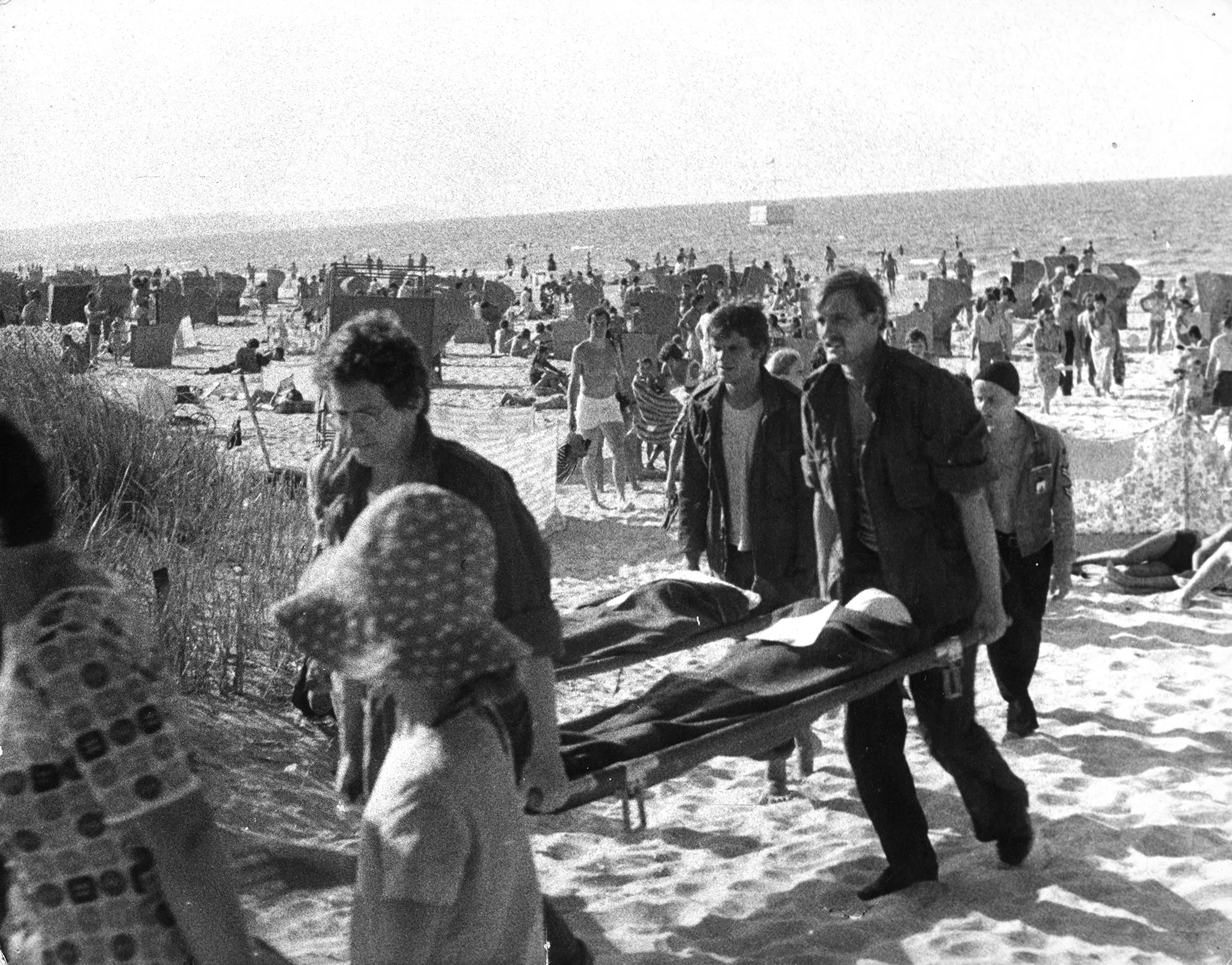 Plaża. Tłum plażowiczów chodzi w kostiumach kąpielowych. W tle widać parawany i morze. Na pierwszym planie grupa aktorów niesie dwie noszy. Nosze wyglądają, jakby leżał na nich ktoś przykryty kocem. Aktorzy niosący nosze ubrani są w jeansowe spodnie i jeansowe kurtki.