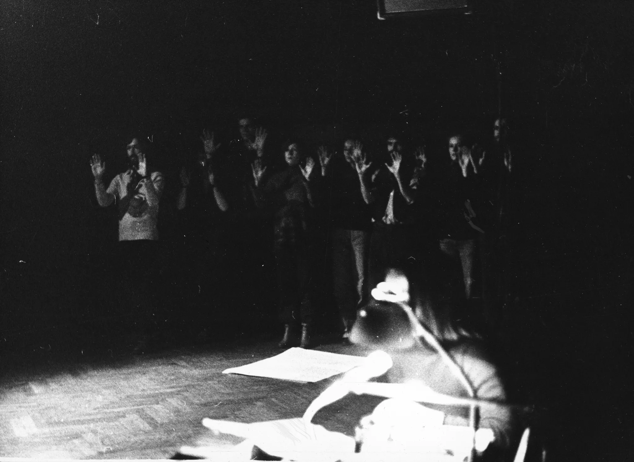 Na drugim planie stoi grupa aktorów i aktorek ustawionych w szregu. Trzymają uniesione, zgięte w łokciach ręce wnętrzem dłoni w stronę widza. Na pierwszym planie niewyraźna postać aktorki siedzącej przy biurku. Oświetla ją lampka.