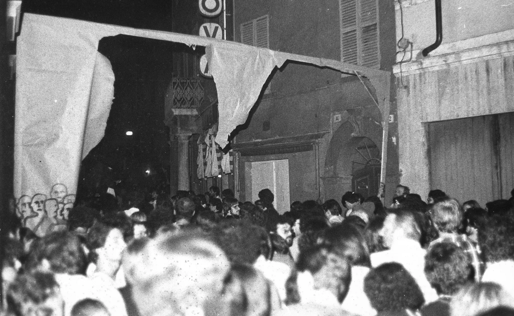 Przestrzeń miejska. Wśród budynków stoi tłum ludzi. Nad tłum stoi prowizoryczna brama. Z lewej strony bramy namalowane są ludzkie sylwetki.