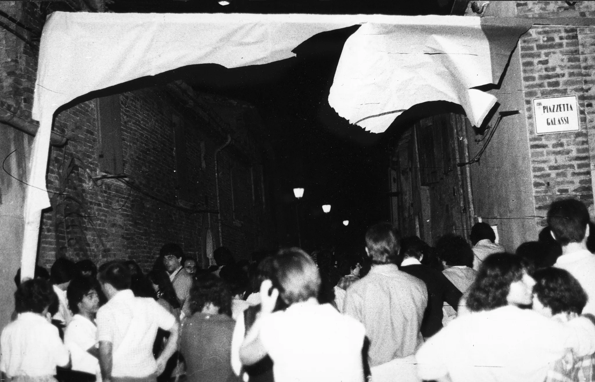 Przestrzeń miejska. Wąską uliczką pomiędzy budynkami przechodzi tłum ludzi. Nad ich głowami zwisają strzępy płachty. Po prawej stronie na ścianie wisi tabliczka z napisem "PIAZETTA GALASSI"