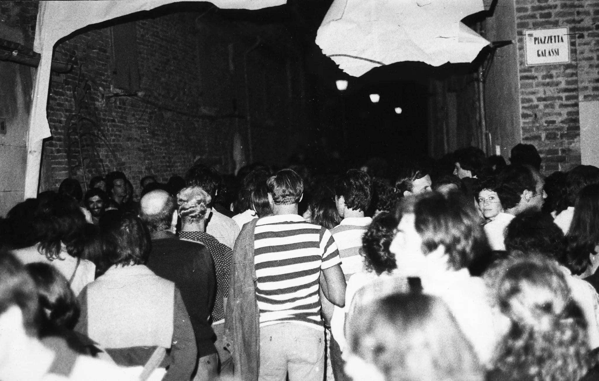 Przestrzeń miejska. Wąską uliczką pomiędzy budynkami przechodzi tłum ludzi. Nad ich głowami zwisają strzępy płachty. Po prawej stronie na ścianie wisi tabliczka z napisem "PIAZETTA GALASSI"