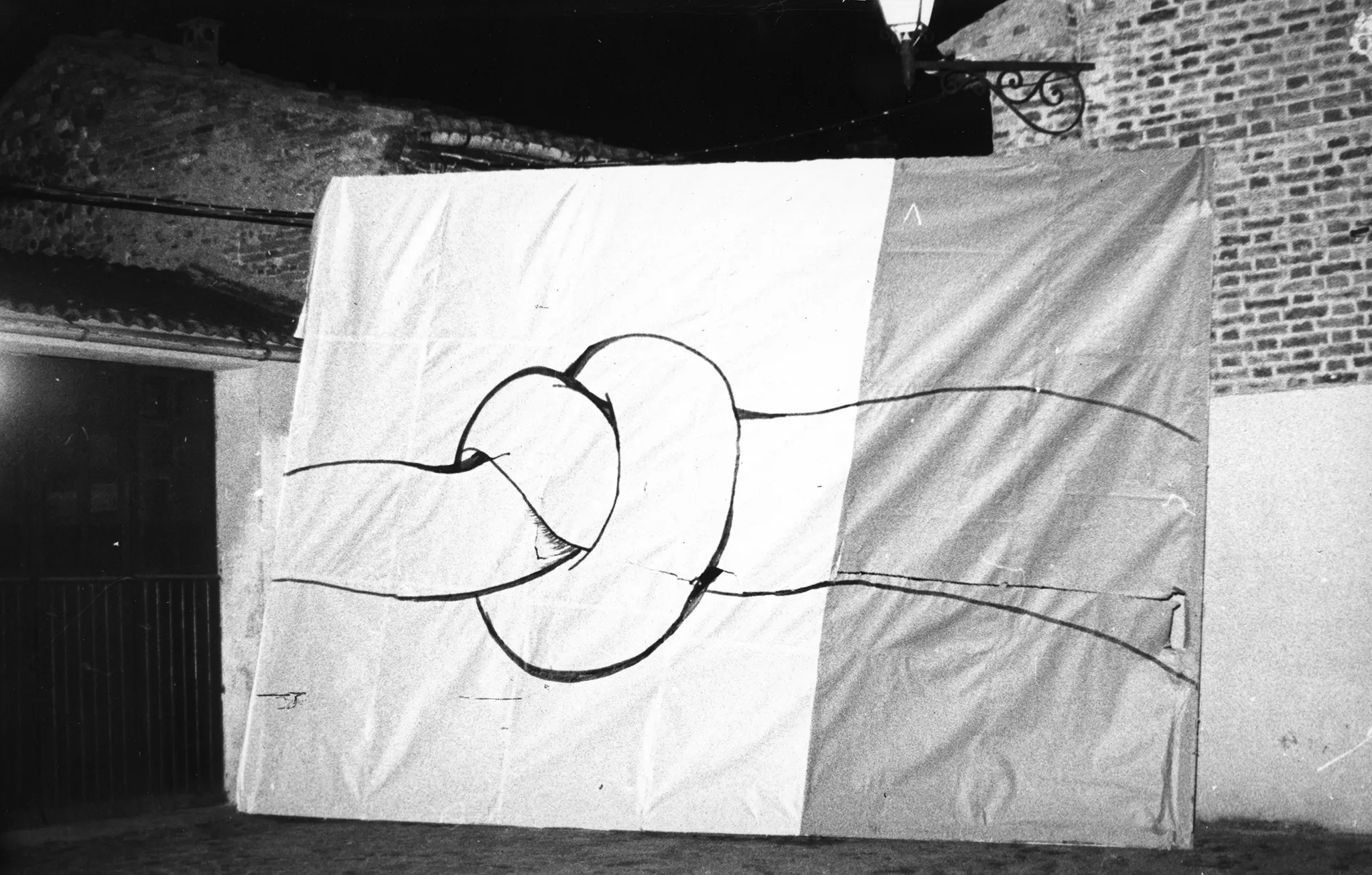 Pod budynkiem stoi wielki biało-szary obraz. Obraz przedstawia spleciony sznur w miejscu splotu.