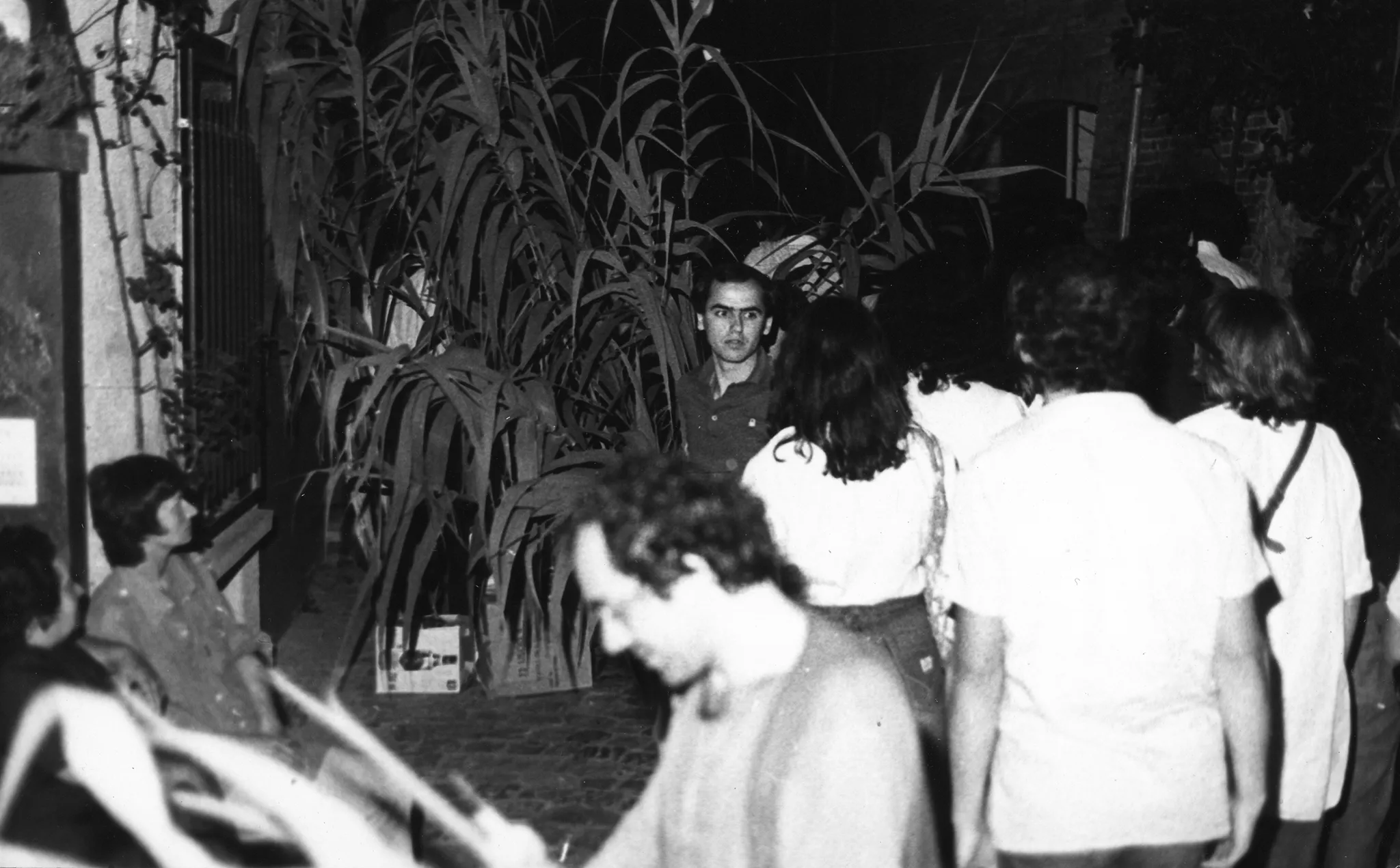 Przestrzeń miejska. Na chodniku stoi grupa osób zwróconych plecami do widza. Sa ubrane na biało. Obok nich, twarzą w stronę widza, stoi młody mężczyzna. Ma na sobie ciemną koszulę. Na pierwszym planie widać zwróconego profilem do widza mężczyznę, który coś czyta. Po lewej stronie na ławce siedzi młoda kobieta. W tle widać wiele roślin.