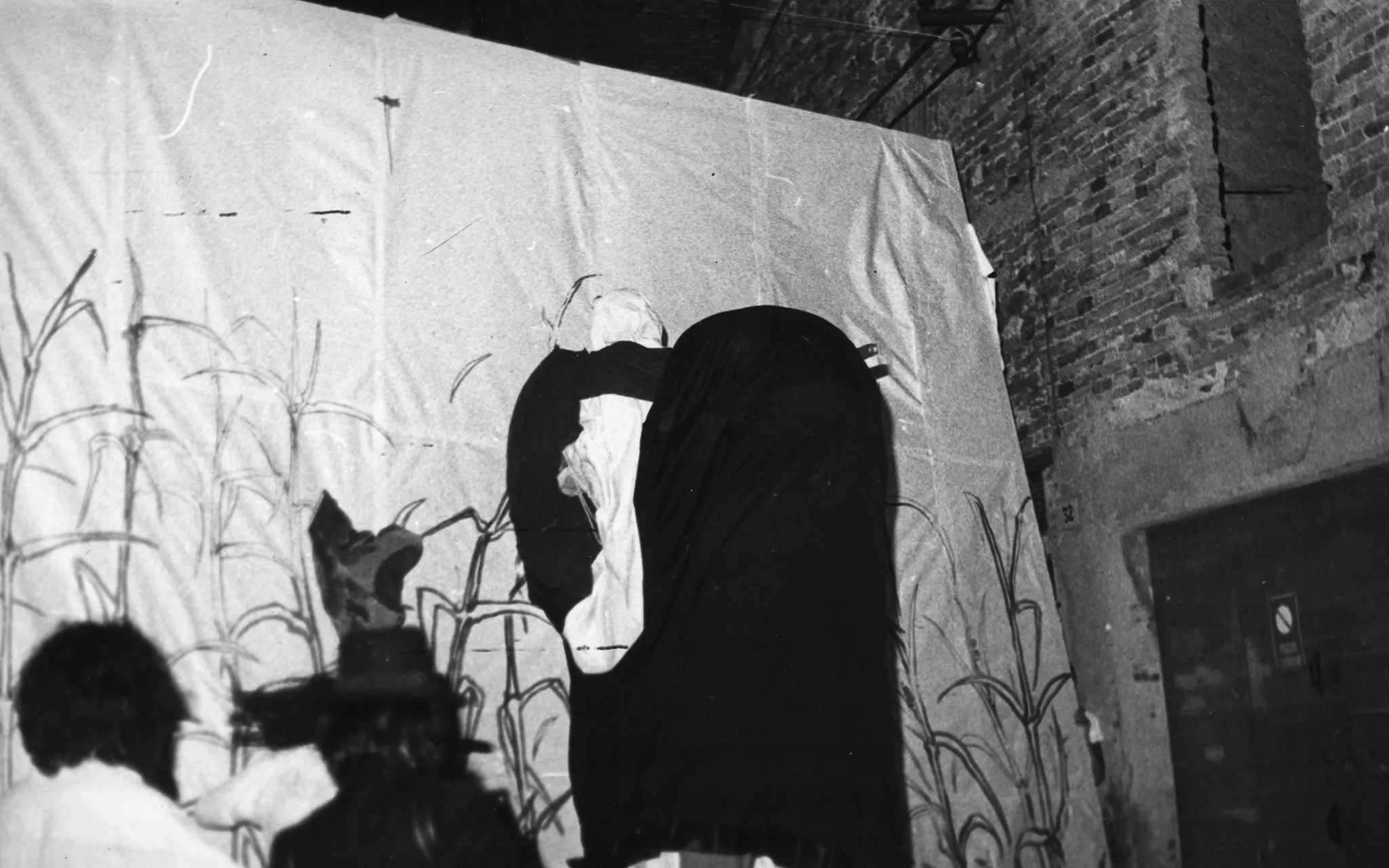 Obok ceglanego budynku stoi wielka biała płachta. Na płachcie namalowane są rośliny przypominające trzciny. Płachta w centrum przysłonięta jest czarnym obłym materiałem. Po lewej stronie stoją dwie osoby zwrócone plecami do widza. Widać tylko ich głowy.