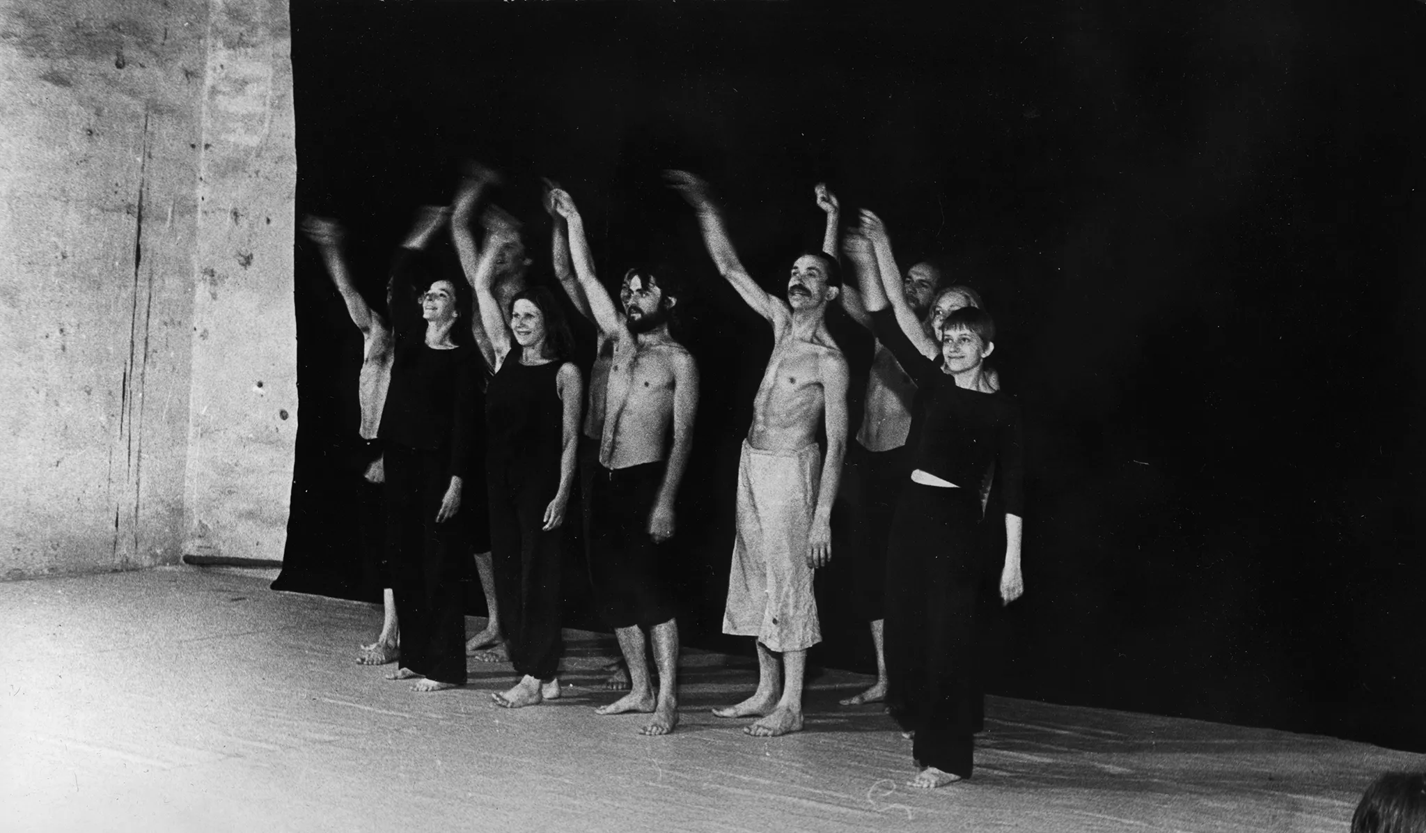 Grupa aktorów i aktorek stoi na tle czarnej płachty. Każdy energicznie macha podniesioną wysoko ręką. Wszyscy są boso. Aktorki ubrane są w czarne spodnie i czarne koszulki. Aktorzy ubrani są w czarne spodenki, mają nagie torsy. Wyróżnia się jeden aktor ubrany w jasne spodenki.