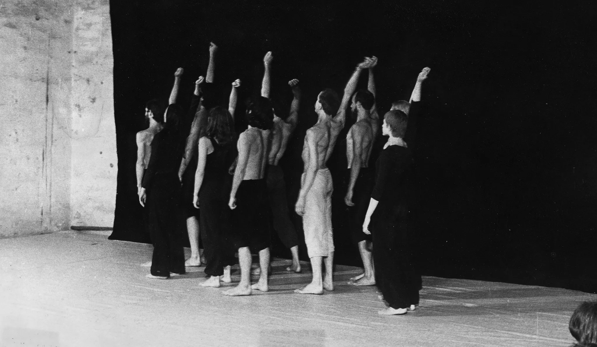 Grupa aktorów i aktorek stoi na tle czarnej płachty. Każdy energicznie macha podniesioną wysoko ręką. Wszyscy są boso. Aktorki ubrane są w czarne spodnie i czarne koszulki. Aktorzy ubrani są w czarne spodenki, mają nagie torsy. Wyróżnia się jeden aktor ubrany w jasne spodenki.