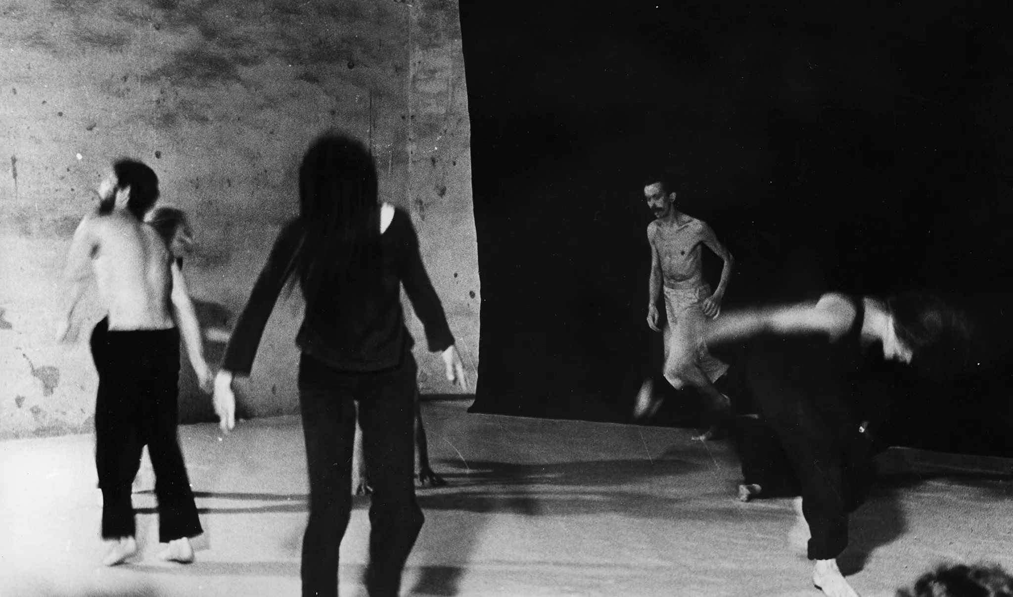 Rozmazane zdjęcie. Tańcząca grupa aktorów i aktorek rozstawionych na sali na tle czarnej płachty. Aktorki ubrane są w czarne spodnie i czarne koszulki. Aktorzy ubrani są w czarne spodenki, mają nagie torsy. Pod czarną płachtą tańczy aktor z nagim torsem ubrany w jasne spodenki. Jest bardzo szczupły.