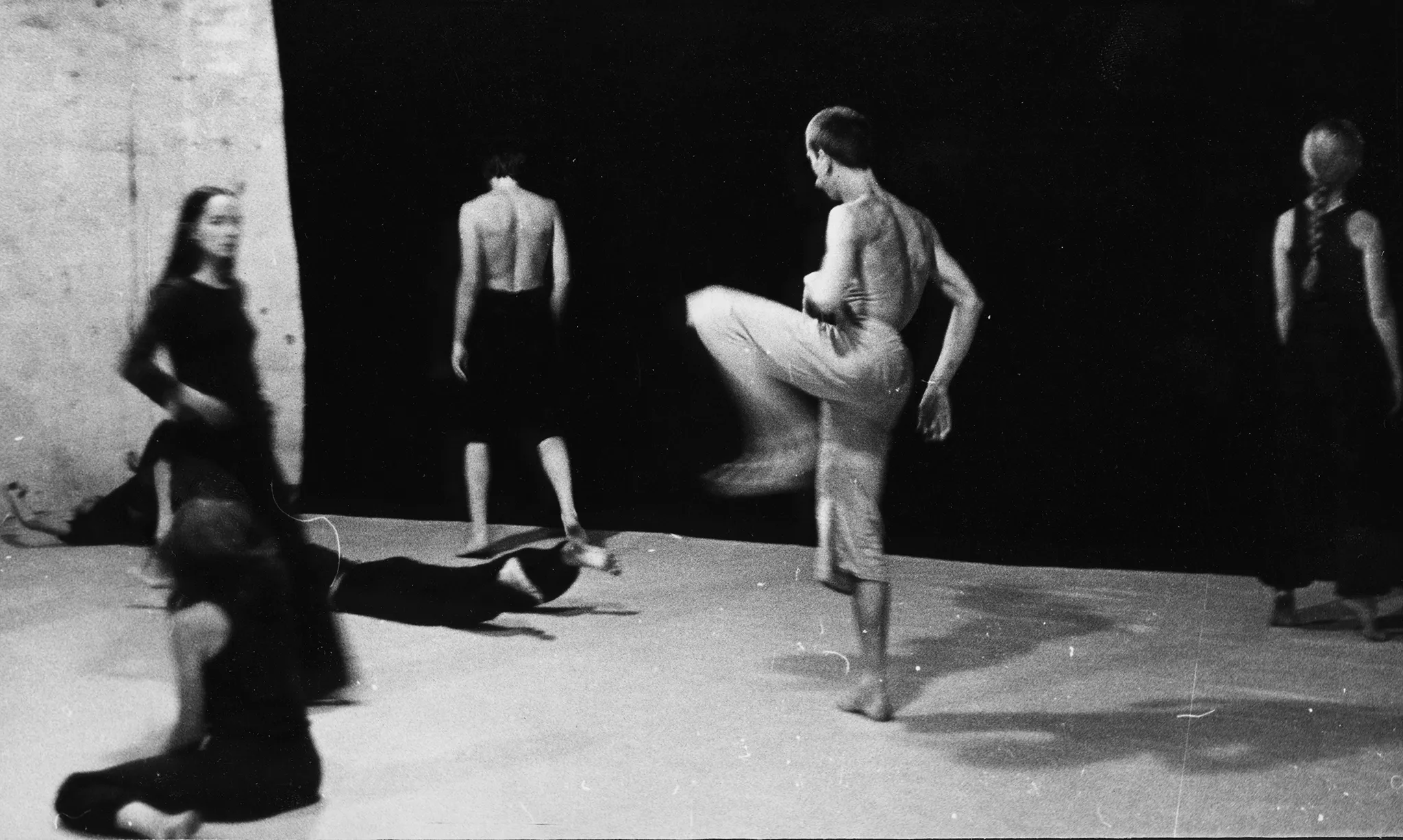 Rozmazane zdjęcie. Grupa aktorów i aktorek rozstawionych na sali na tle czarnej płachty. Po lewej stronie nieruchome aktorki ubrane na czarno. W centrum na jednej nodze odwrócony plecami do widza stoi aktor ubrany w jasne spodenki. Jego tors jest nagi. Po dwóch bokach czarnej płachty stoi aktor i aktorka. Są odwróceni plecami w stronę widza. Aktor ubrany jest w czarne spodenki, ma nagi tors. Aktorka ubrana jest w czarne spodnie i czarną koszulkę na ramiączka. Ma włosy zaplecione w warkocz.