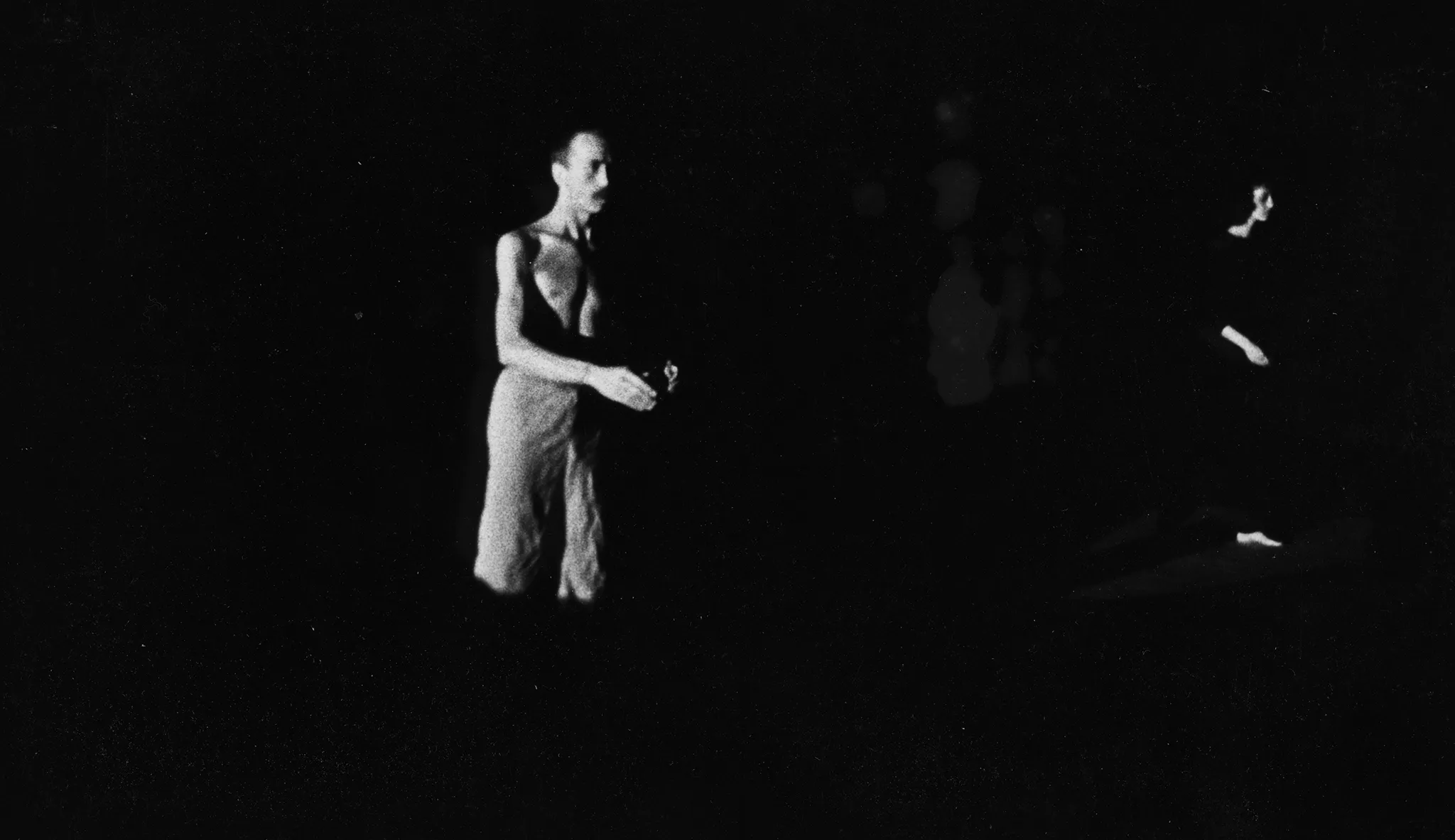 Niewyraźne zdjęcie. Z cienia wyłania się aktor stojący profilem w stronę widza. Ubrany jest w jasne spodenki, jego tors jest nagi. Po prawej stronie stoi słabo widoczna aktorka. Ubrana jest na czarno.