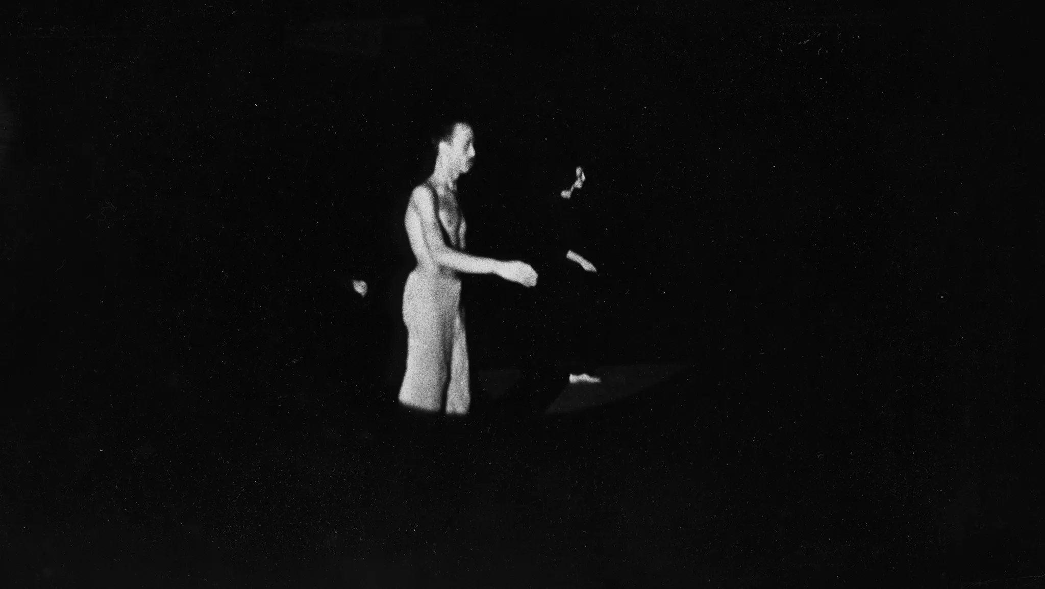 Niewyraźne zdjęcie. W świietle reflektora stoi aktor zwrócony profilem w stronę widza. Ubrany jest w jasne spodenki, ma nagi tors. Delikatnie unosi jedną rękę. Za nim stoi aktorka ubrana na czarno. Przybiera taką samą pozę jak aktor.