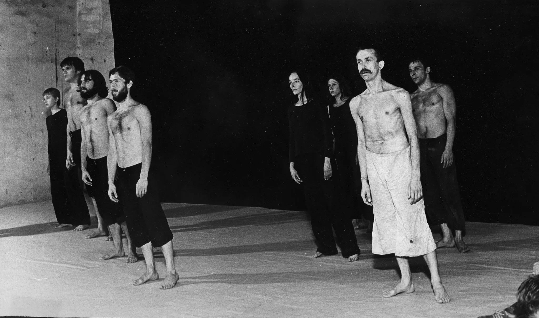Grupa aktorów i aktorek stojących na tle czarnej płachty. Aktorzy ubrani są w czarne spodenki, mają nagie torsy. Aktorki ubrane są w czarne spodnie i czarne koszulki. Wśród grupy wyróżnia się aktor ubrany w jasne spodenki. Wszyscy są boso. Wszyscy delikatnie przechylają się w tę samą stronę.