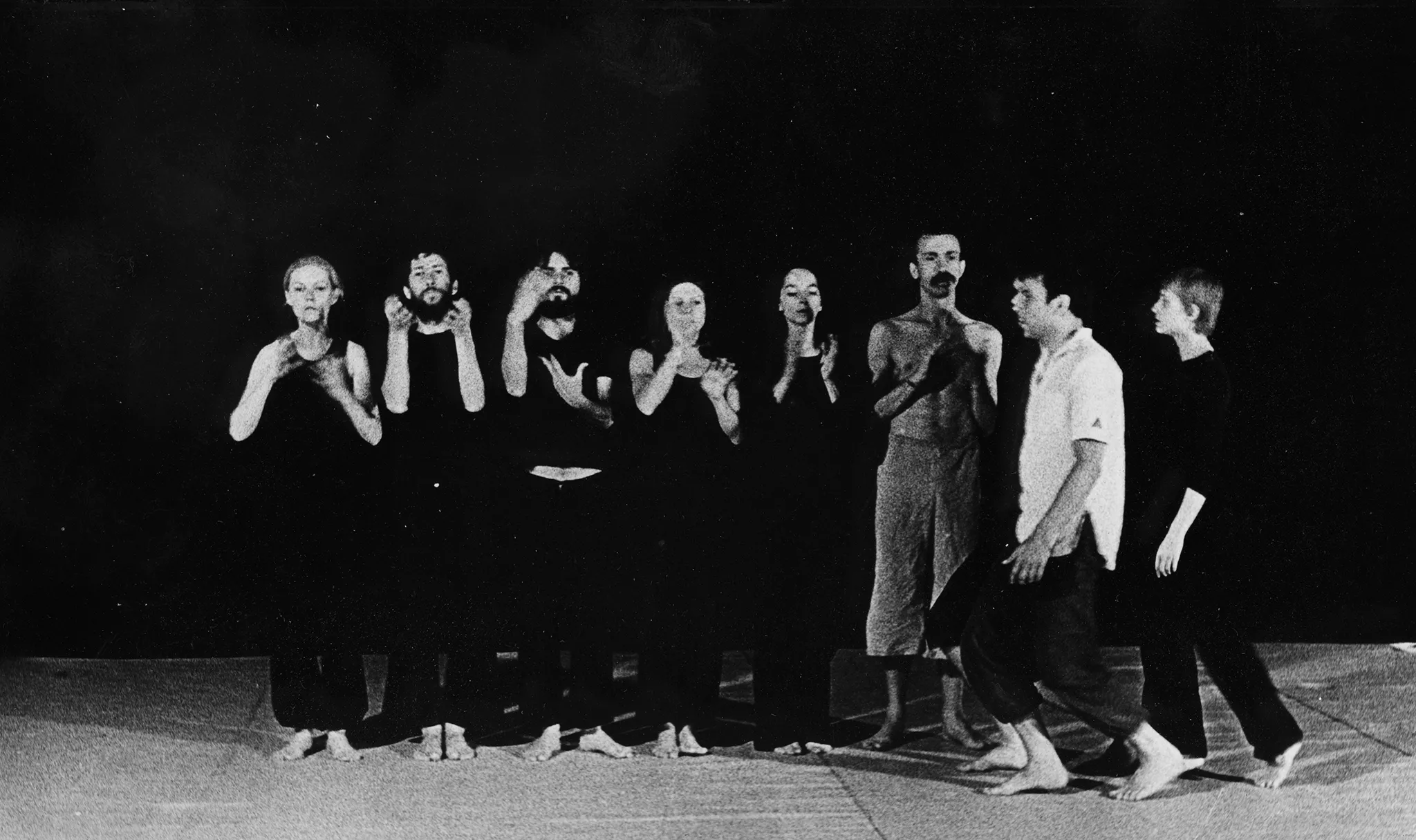 Grupa aktorów i aktorek stojących w szeregu na tle czarnej płachty. Wszyscy są boso i ubrani w czarne ubrania. Wyróżnia się aktor ubrany w jasne spodenki. Z prawej strony idzie grupa aktorów i aktorek zwróconych profilem w stronę widza.